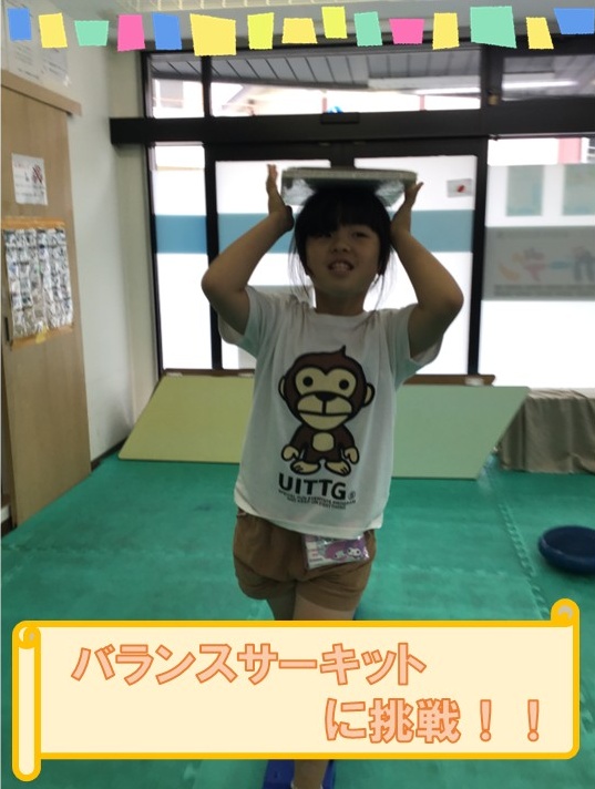 Child balances a tray on her head in a playroom, wearing a white monkey T‑shirt; banner in Japanese reads 'バランスサーキット に挑戦！！'