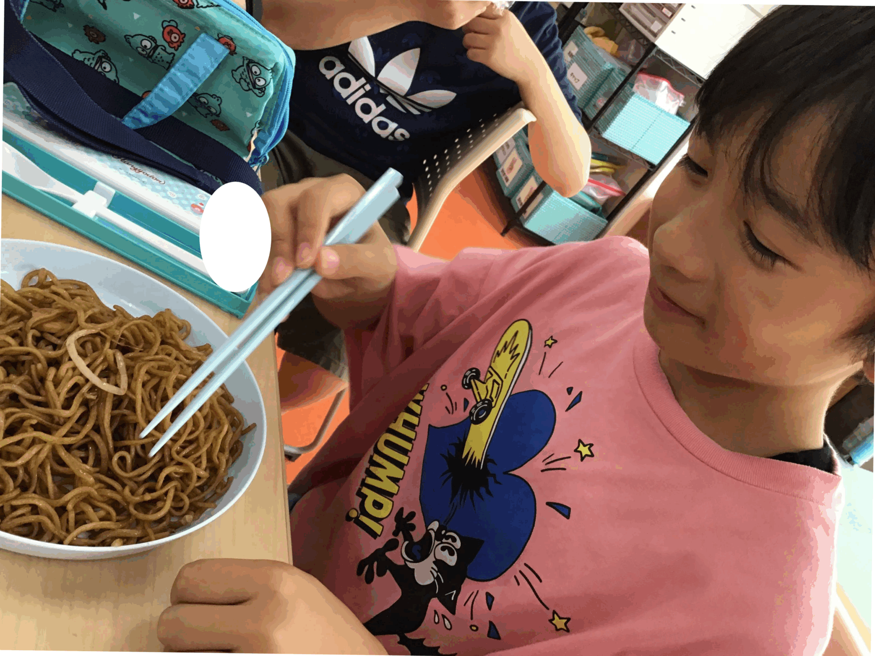 Child in a pink shirt uses blue chopsticks to pick up noodles from a bowl at a cafeteria table