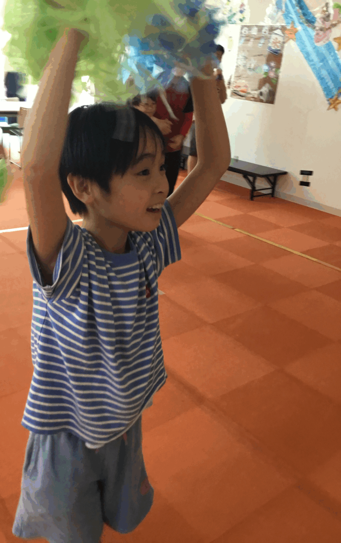 Smiling child in a blue striped shirt raises arms to hold colorful streamers indoors with decorations on the wall behind him.