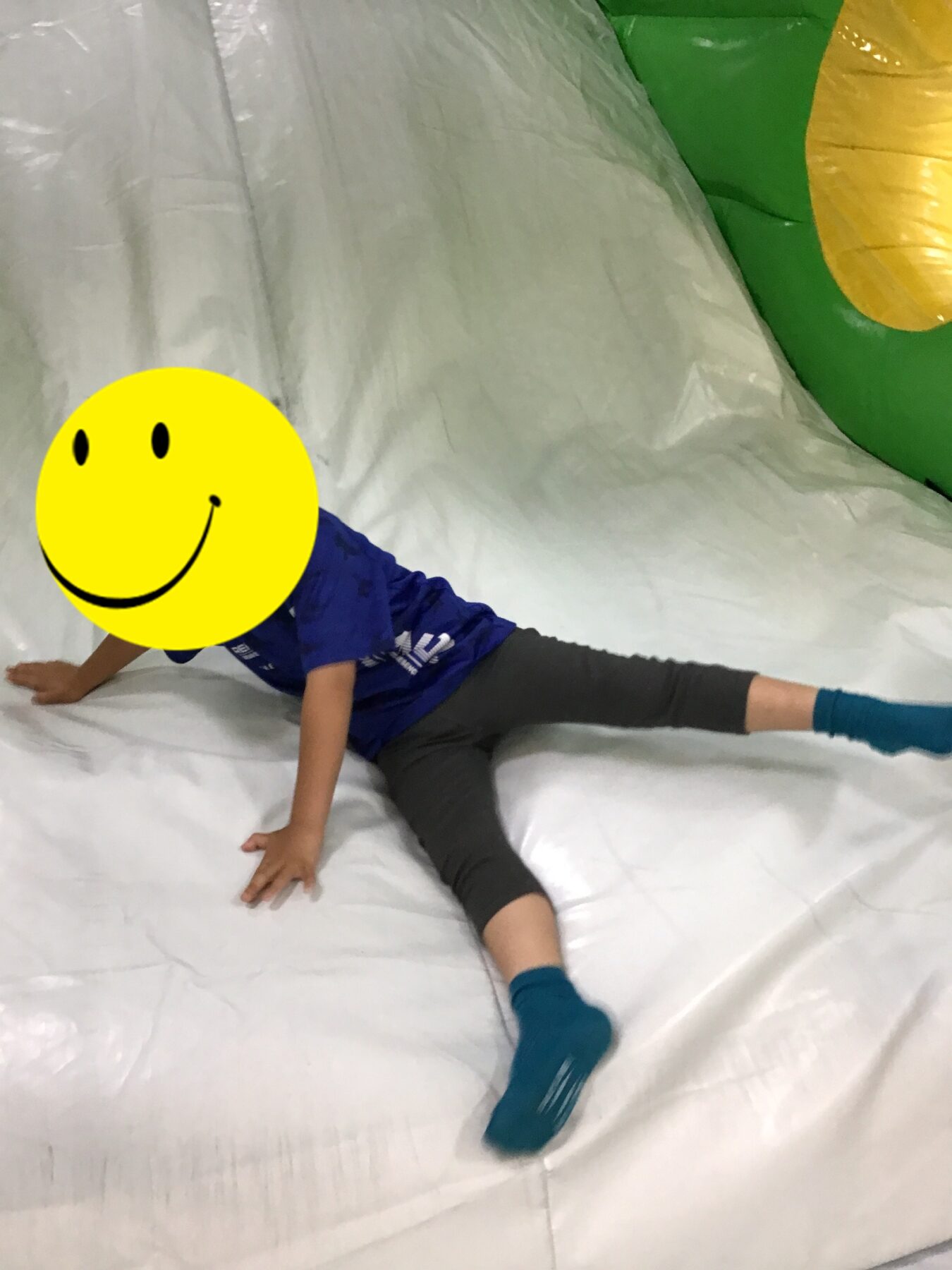 Child sliding or climbing on an inflatable indoor play area, wearing teal socks and gray leggings, with a yellow smiley face covering their face for privacy.