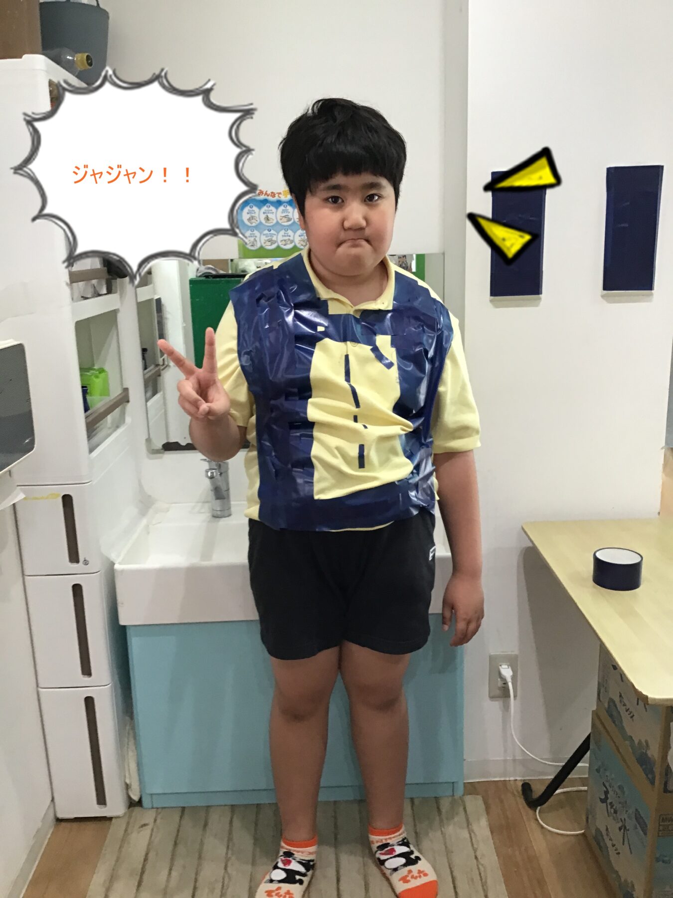 Young boy standing in a room wearing a blue duct-tape vest over a yellow shirt, flashing a peace sign with two fingers.