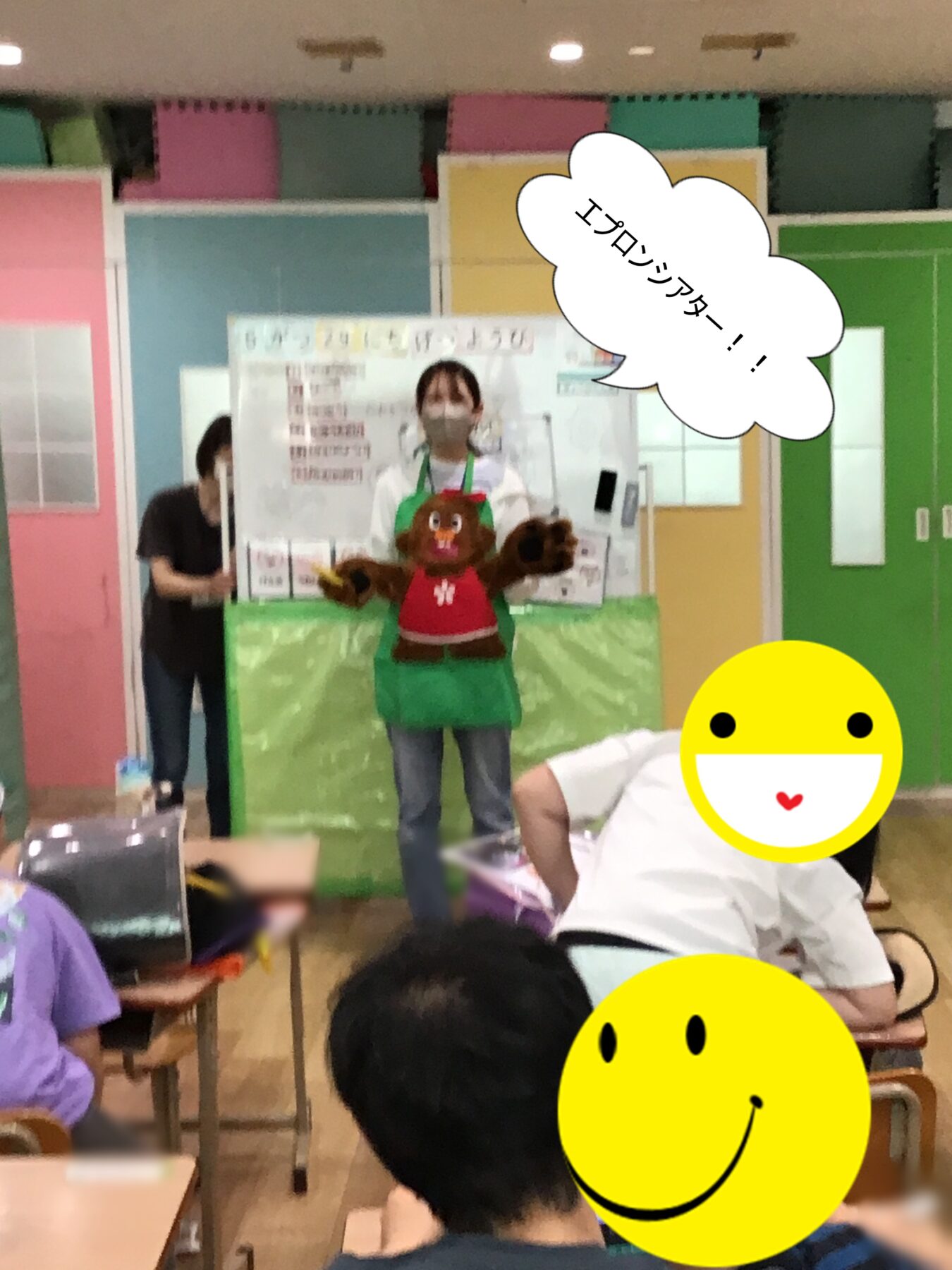 Instructor in a bear-themed apron stands on a small stage in a bright, color-blocked classroom, addressing seated children.