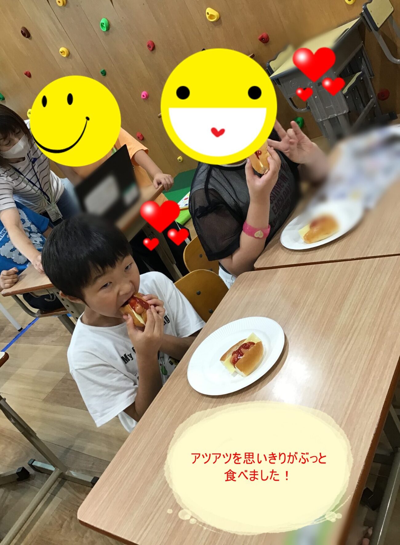 Group of children at a school table eating hot dogs with ketchup, colorful climbing-wall holds on the wall behind, faces covered with large emoji stickers (privacy).
