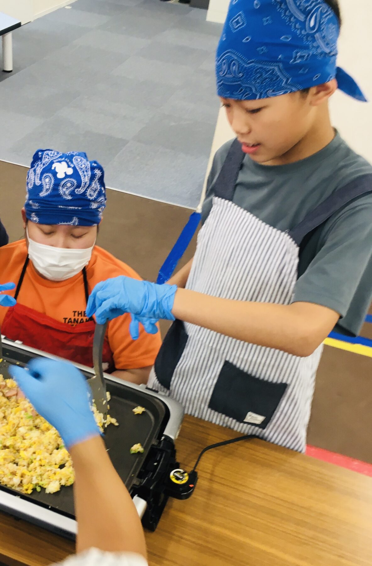 Two kids wearing blue bandanas and aprons cook fried rice in a portable pan on a table, with one stirring the dish.