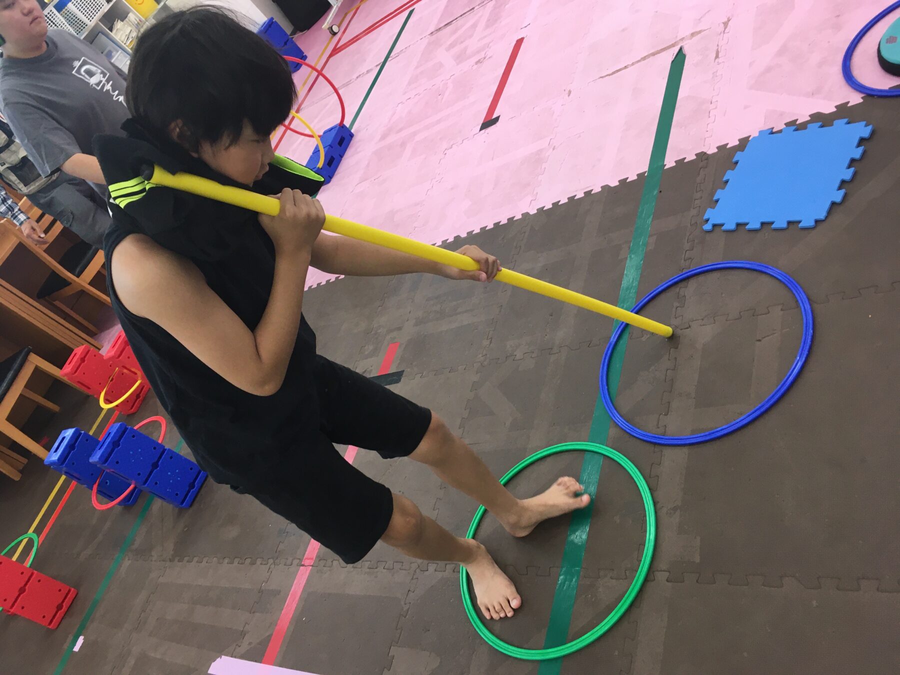 Child holding a yellow baton and guiding it through a green hoop on a padded gym floor.