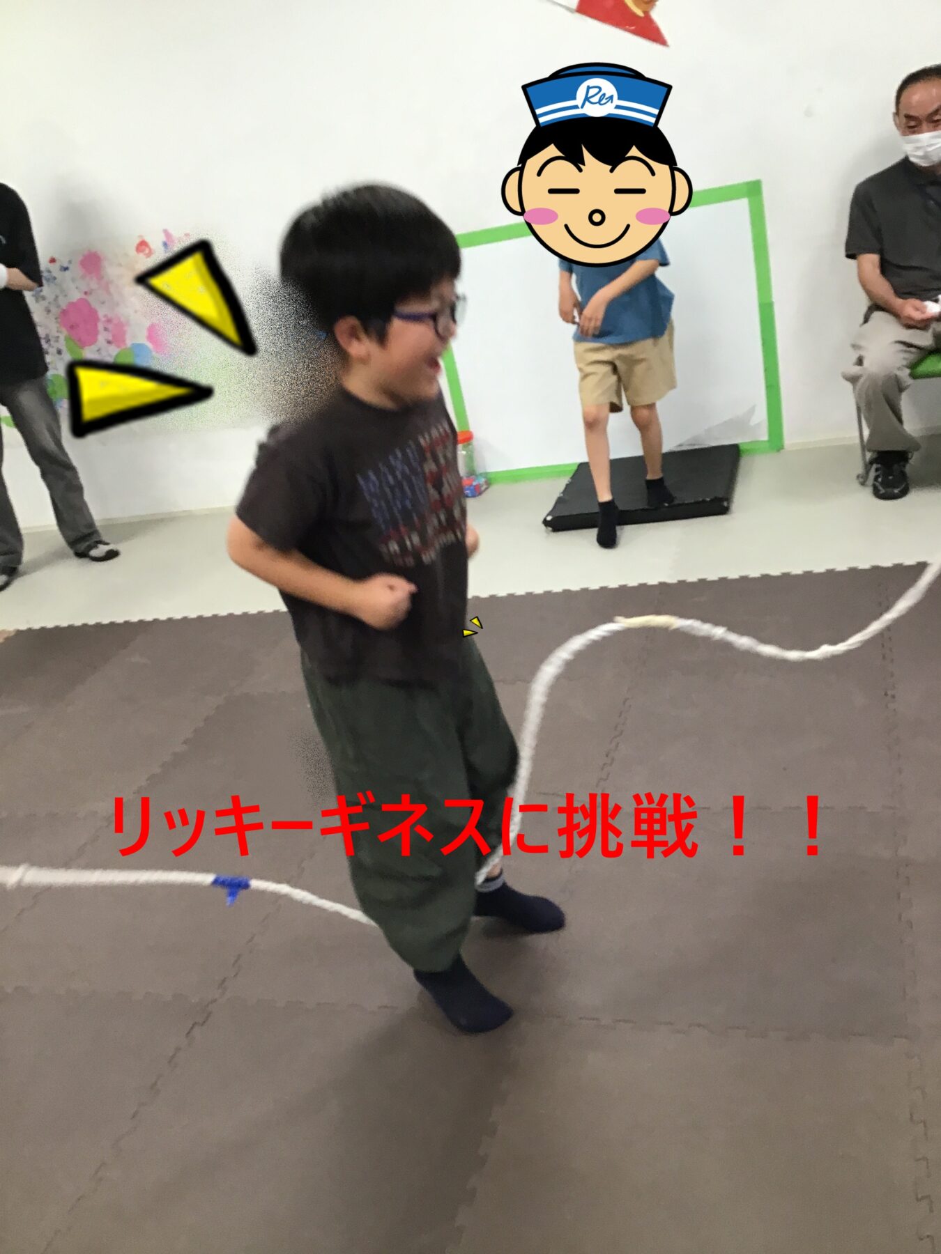 A boy in a dark shirt runs on a padded floor while another child stands on a small black platform; red JP text at bottom reads “リッチーゲネスに挑戦！！” and adults watch in the background.