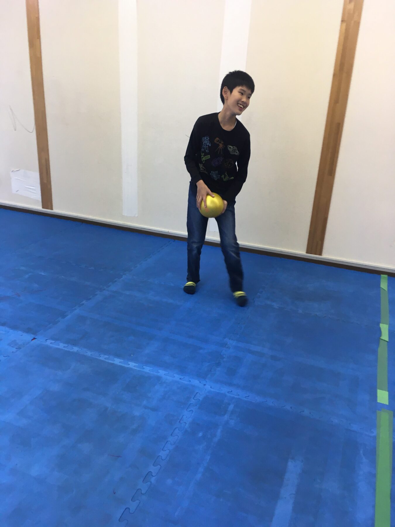 Boy standing on a blue gym mat, smiling while holding a yellow ball with both hands at waist level inside a room with white walls and wooden posts.