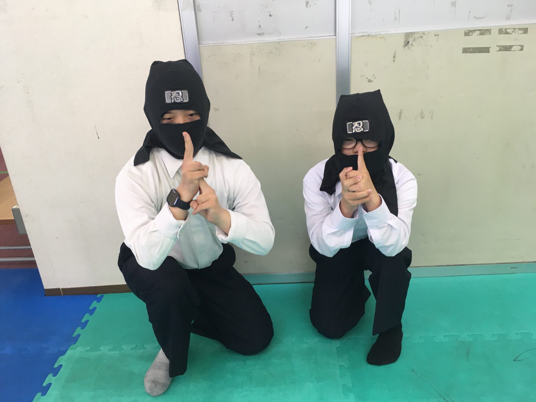 Two people in ninja costumes kneel on a colorful foam mat, making a shushing gesture with a finger to the lips (patch that reads 忍 on their hoods).
