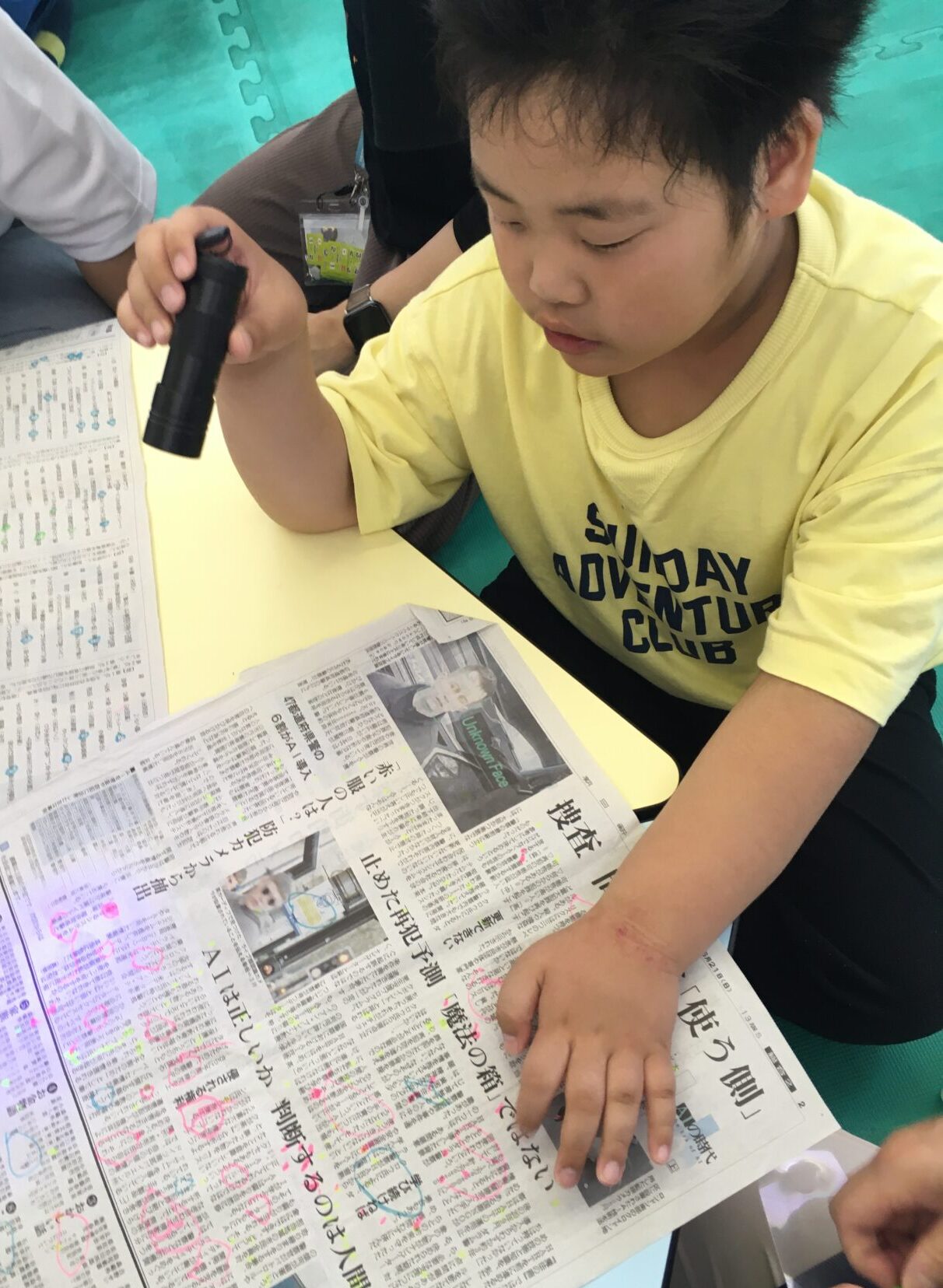 Young boy in a yellow shirt reads and marks a Japanese newspaper with a highlighter at a desk.