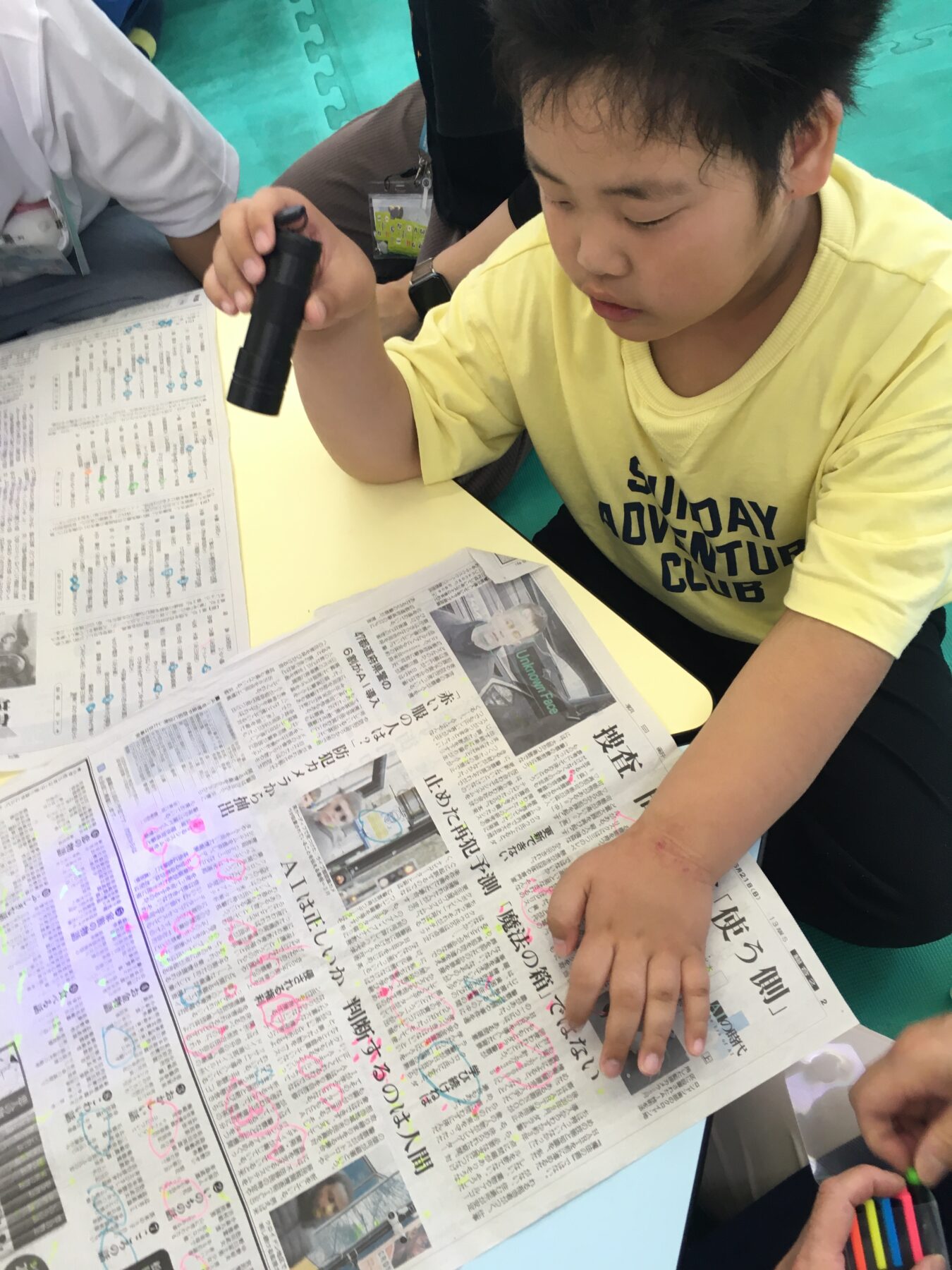Young boy in a yellow shirt reads and marks a Japanese newspaper with a highlighter at a desk.