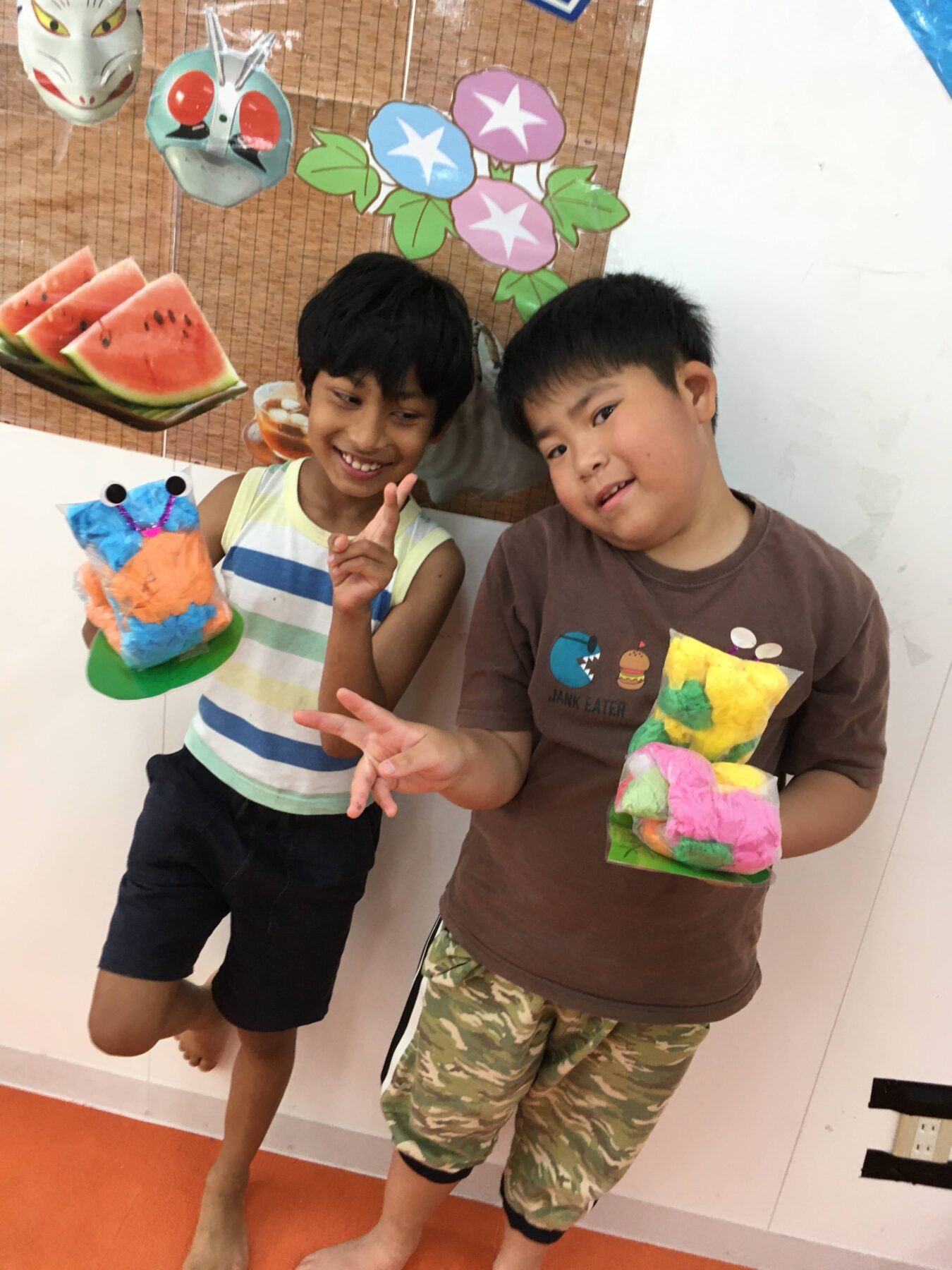 Two smiling boys pose indoors, each holding colorful handmade crafts in front of a fruit-and-flower mural backdrop