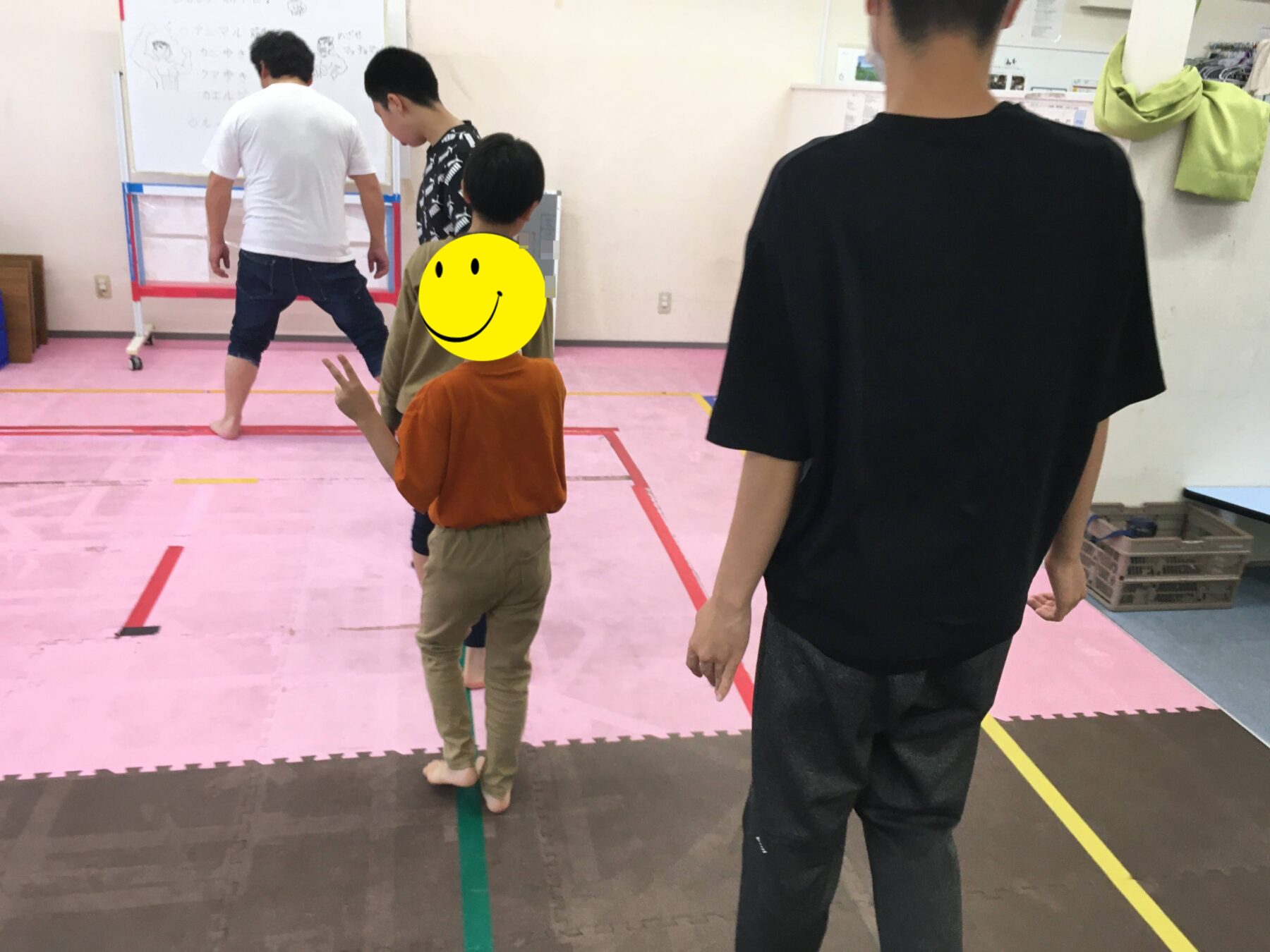 Group of people standing on a pink gym mat, watching a whiteboard at the front of a studio. A child in orange holds up a peace sign, while a person in a white shirt and a person in a patterned shirt look toward the board, and another person in a black shirt stands nearby in the foreground.