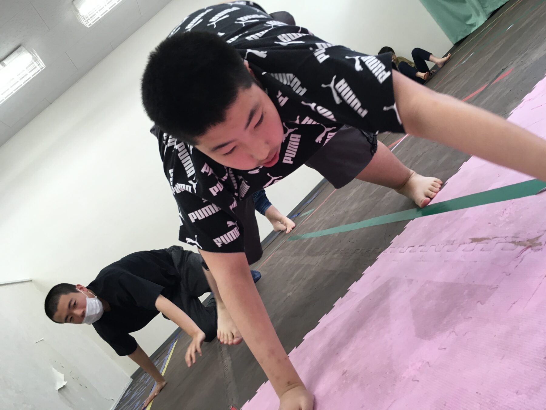 People in a gym on pink interlocking mats doing a forward-reaching exercise; foreground person in a black Puma shirt and shorts leans toward the floor with arms extended.