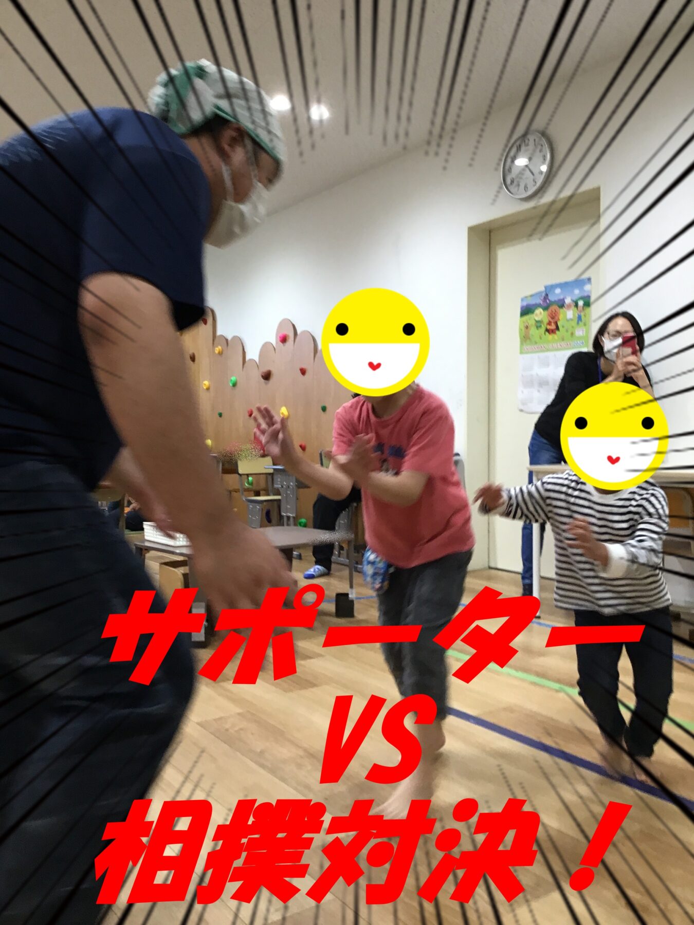 Indoor playroom scene with a man in a mask and green cap leaning toward running children; a boy in pink reaches forward and a child in stripes runs nearby, both with emoji faces covering their features. A caregiver watches and shoots with a phone in the background; colorful climbing holds and a wall are visible.