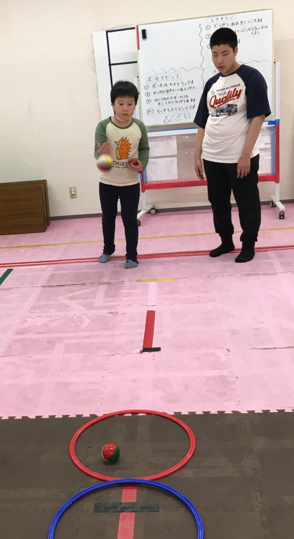 Young boy throws a ball while a man watches in a gym, with red and blue hoops placed on the padded floor behind them.