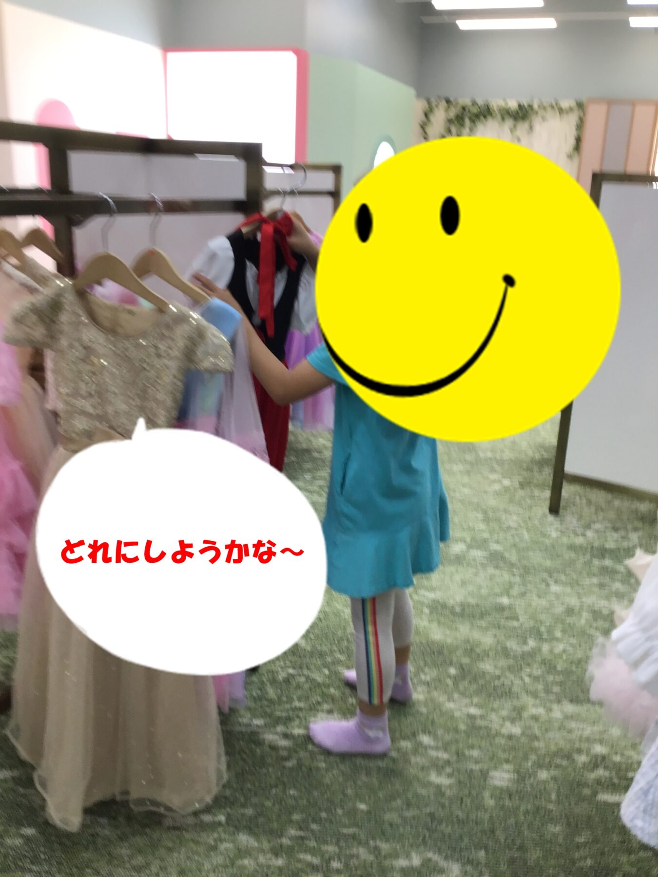 A child in a turquoise dress tries on dresses at a clothing store, browsing a rack of gowns; a large yellow smiley sticker covers their face. A white speech bubble with red Japanese text appears in the foreground.