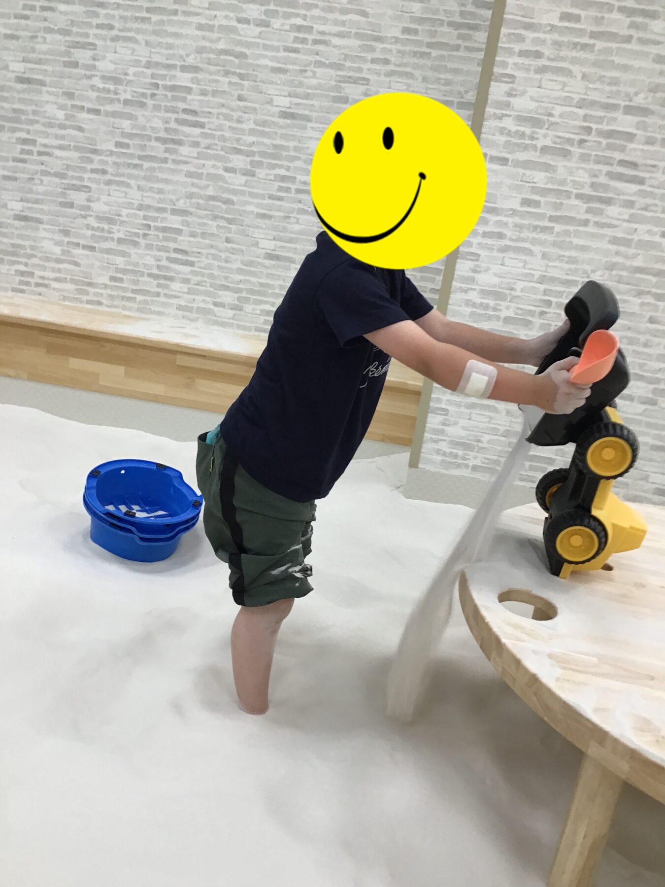 Child in a dark T-shirt and green shorts pushes a yellow and black toy vehicle on a white play surface, with a blue bucket nearby.