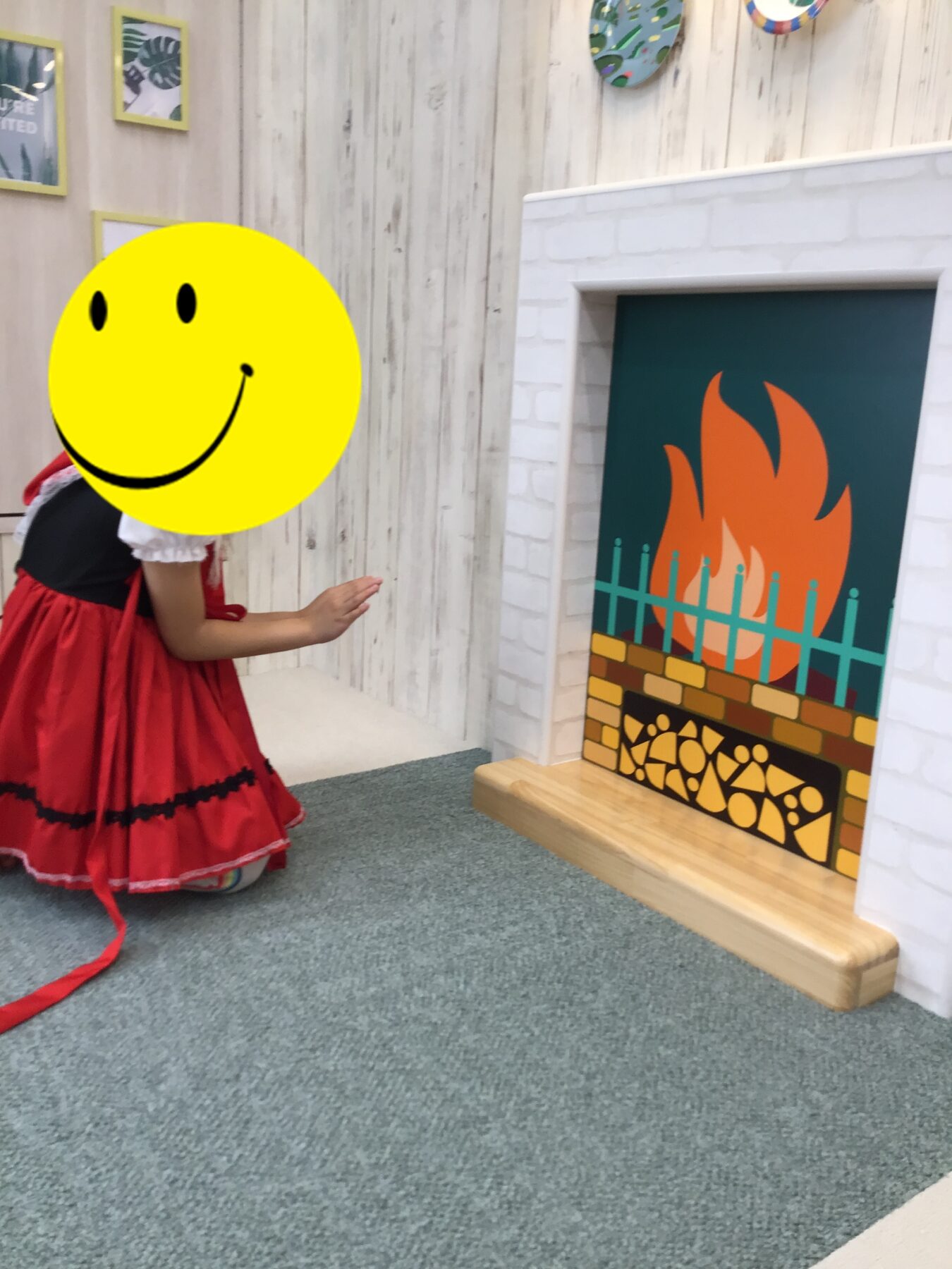 Child in a red dress kneels on a carpeted floor beside a painted fireplace; their head is covered by a large yellow smiling-face graphic.