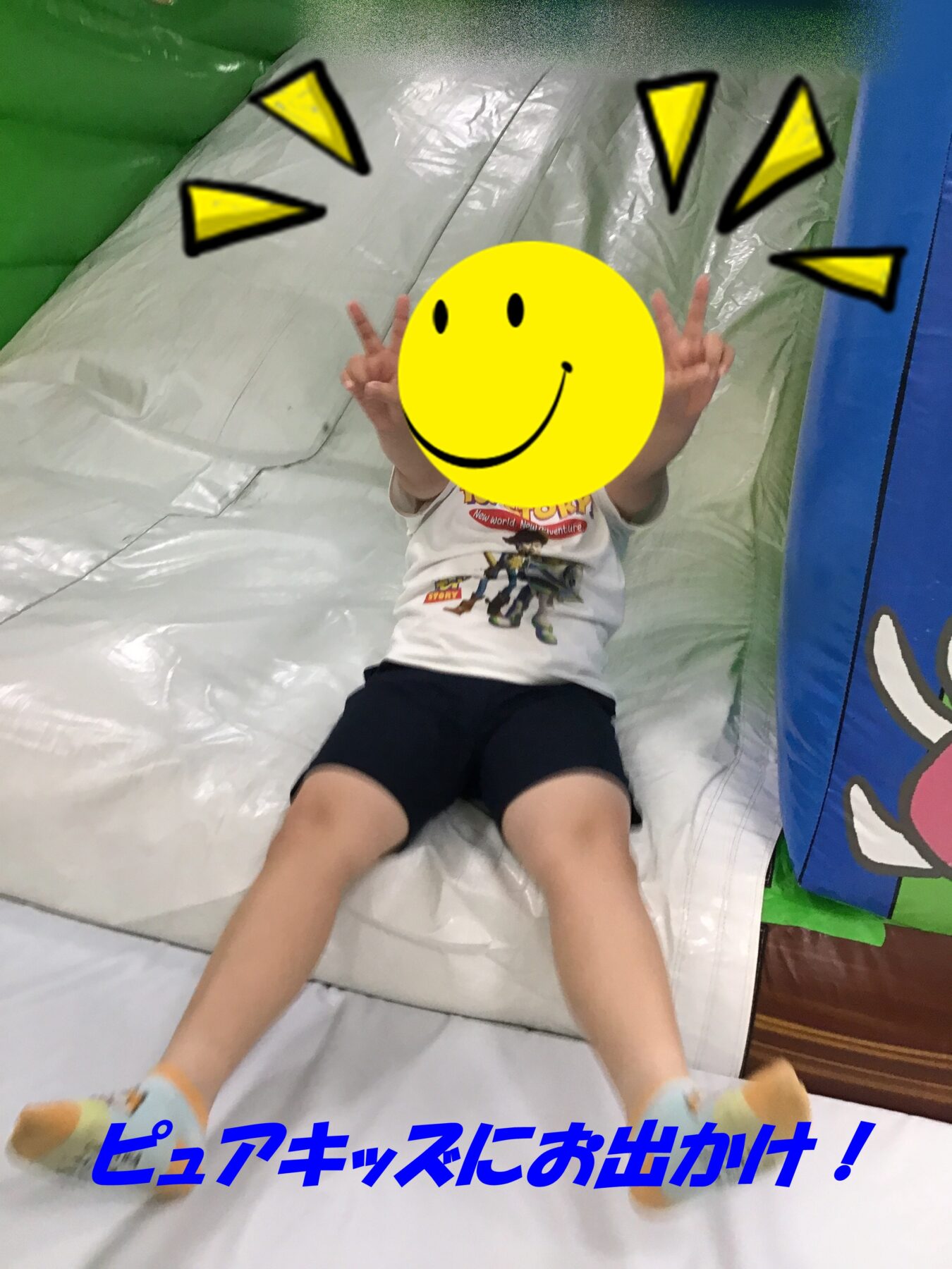 Child sits at the bottom of a white inflatable slide, making peace signs with both hands; yellow smiley sticker covers the face for privacy.