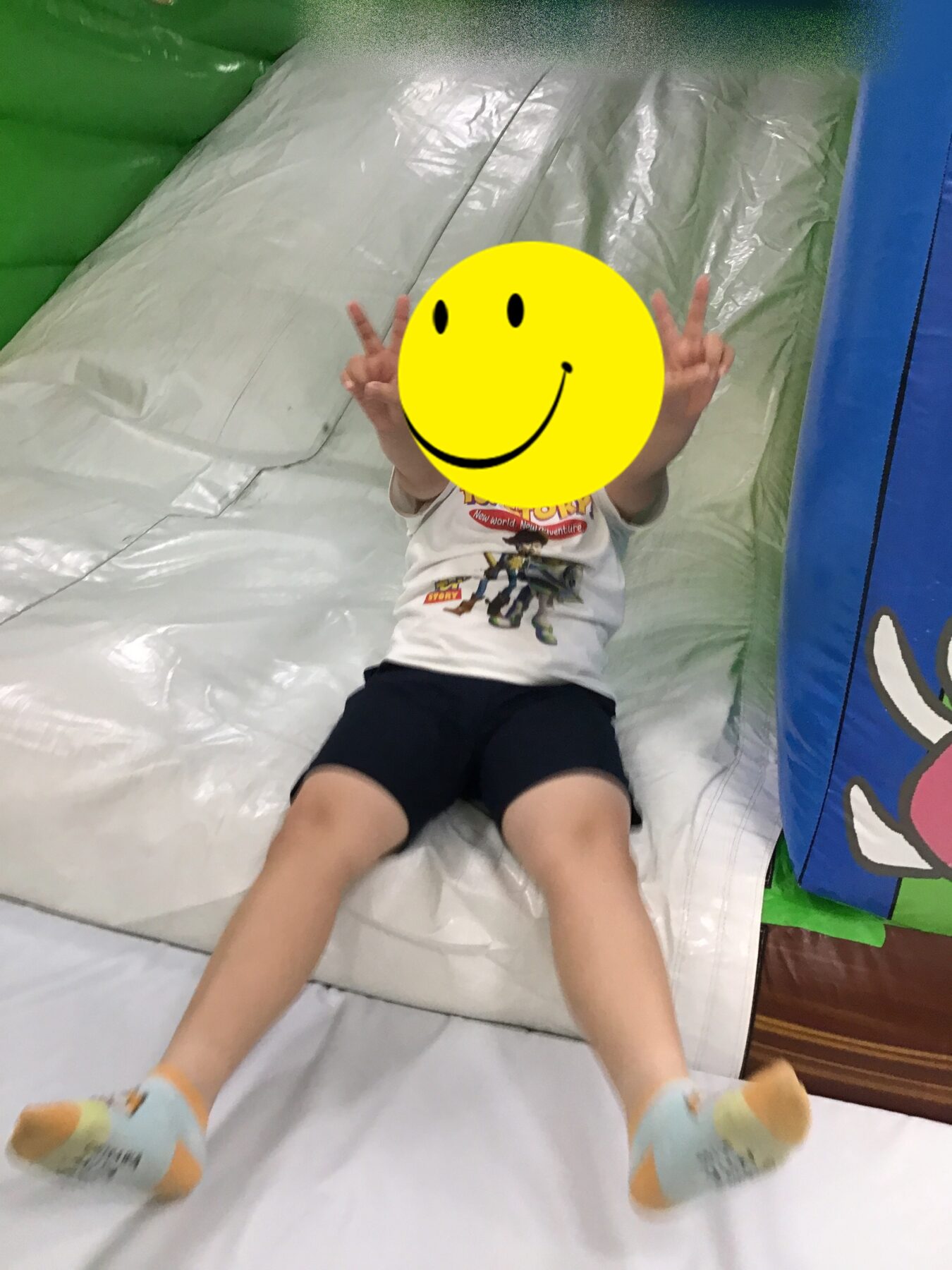 Child on a soft indoor slide, arms raised in peace signs, face covered by a yellow smiley sticker; wearing a white graphic T‑shirt and dark shorts.