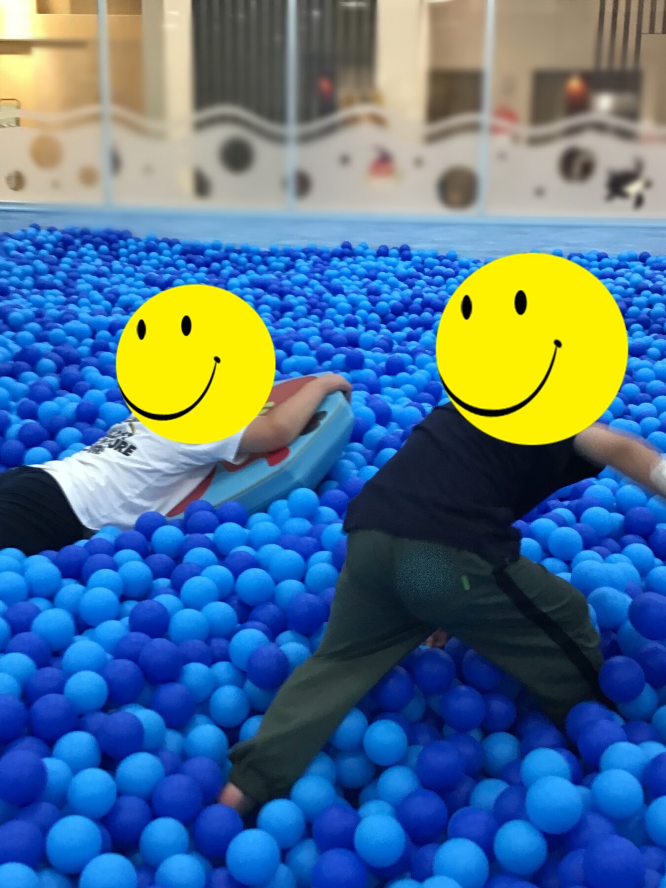 Two children play in a blue ball pit indoors, smiling emoji stickers over their faces as they lean on a floaty boat.