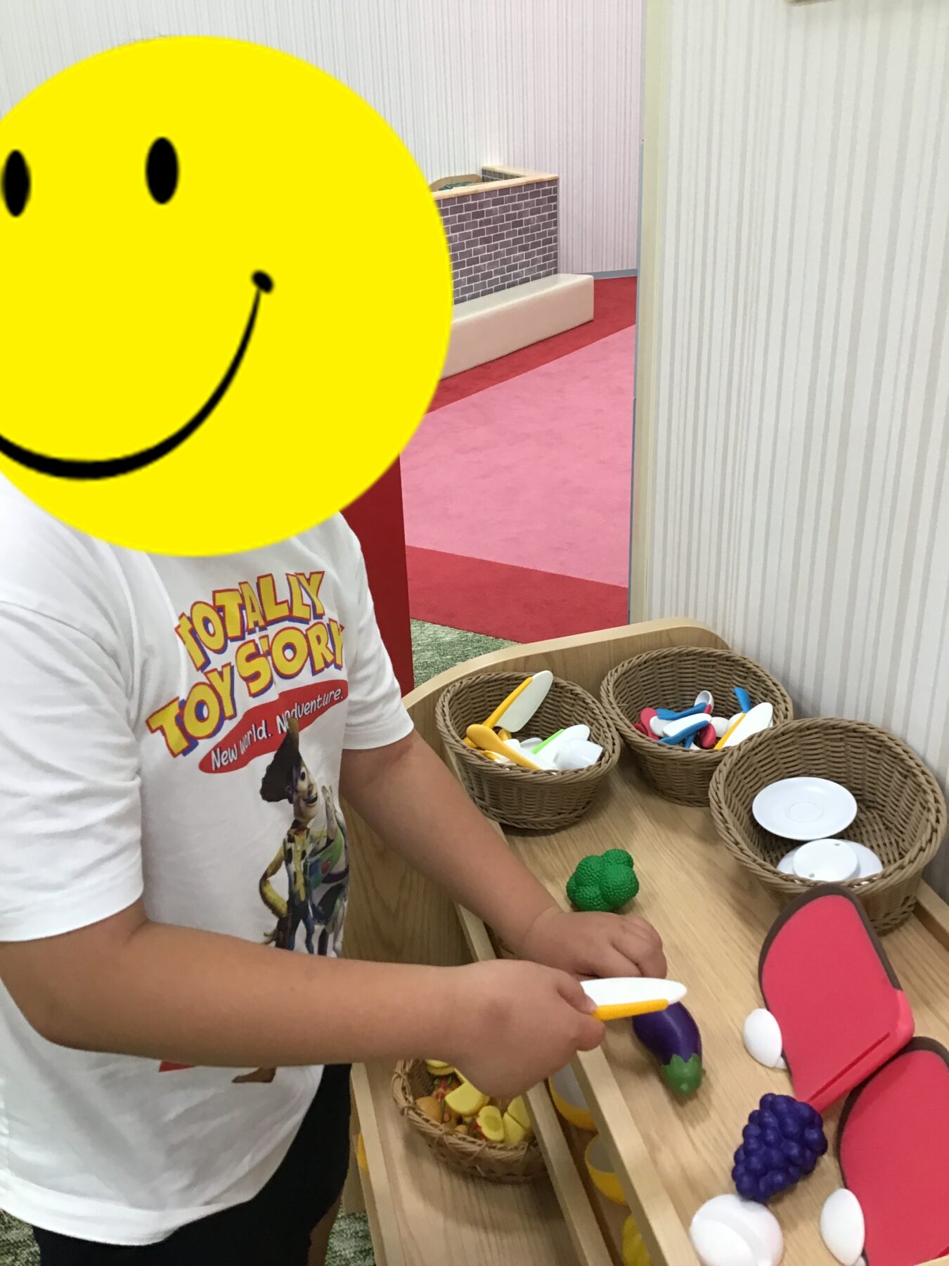 Child playing with a wooden toy kitchen, handling plastic utensils and pretend food at a table; smiley face overlay covers their face.