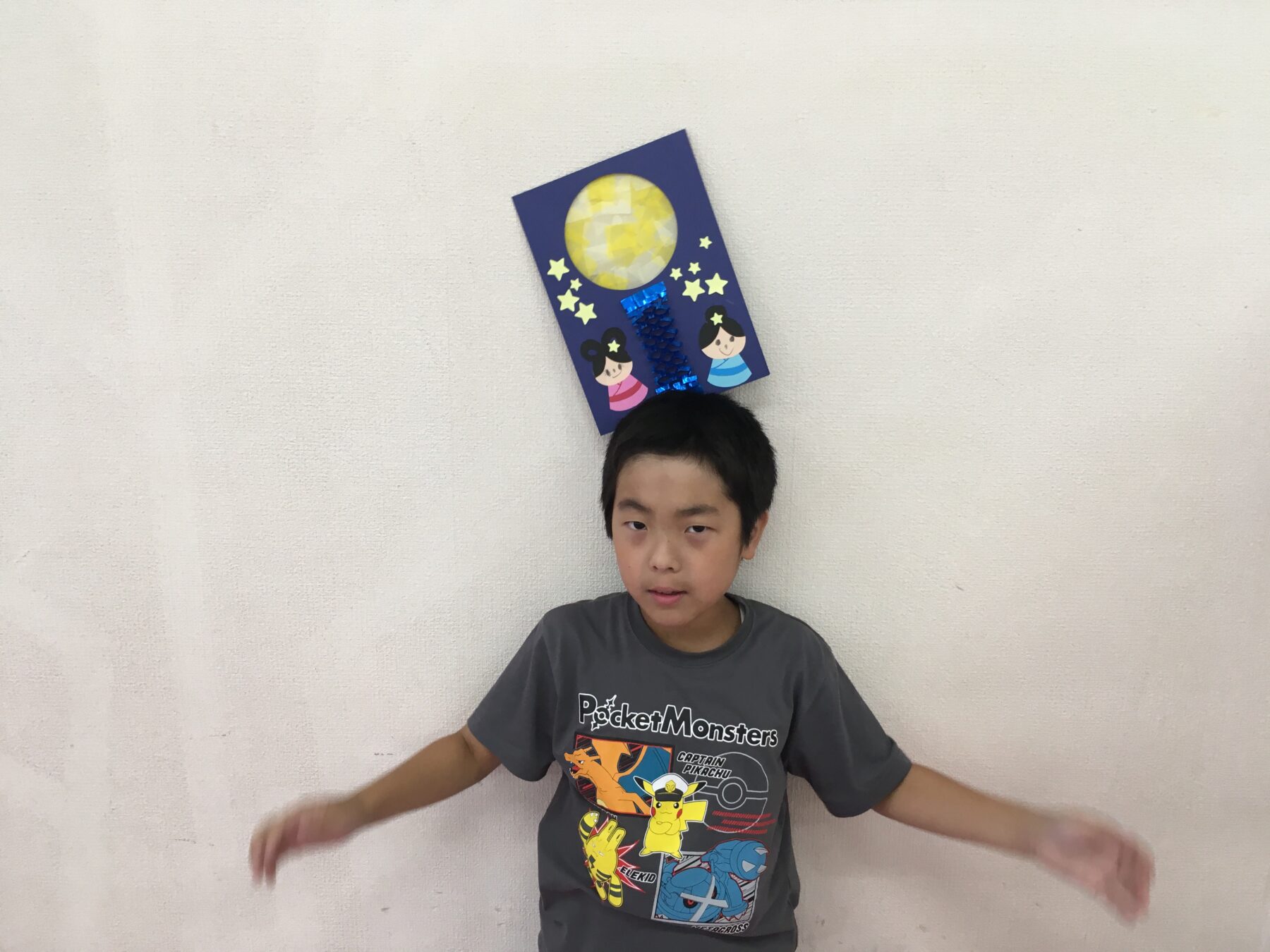 Boy wearing a handmade blue poster with a yellow moon and stars on his head, posing against a light wall; arms outstretched.