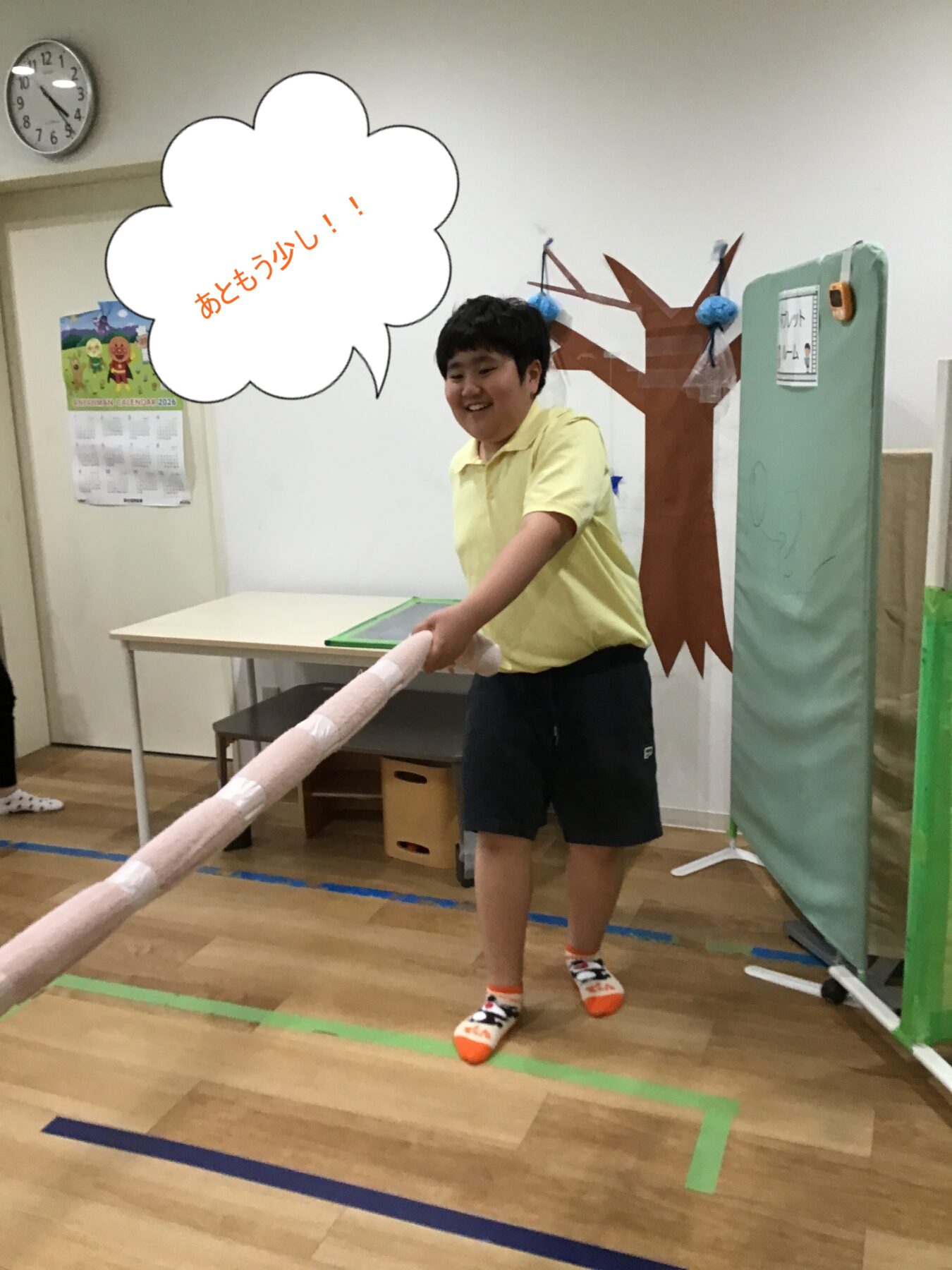 Boy in a yellow polo shirt smiles while holding a long foam baton in a classroom gym, with a paper tree backdrop and foam matting nearby.