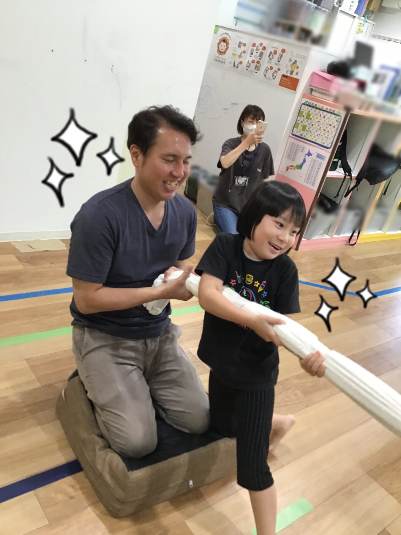 Man kneels on a cushioned block, smiling while a young boy pulls a white towel during a playful tug-of-war in a gym. A woman records with her phone in the background.