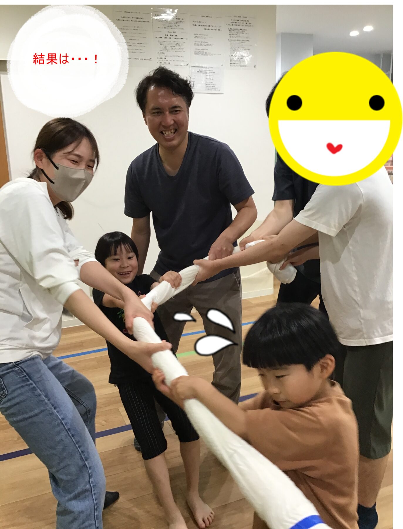 Group of adults and kids pulling a white rope in a gym, smiling and leaning forward as they compete; a large yellow smiley face covers one participant on the right; Japanese text reads '結果は…!' in a white circle at the top left.