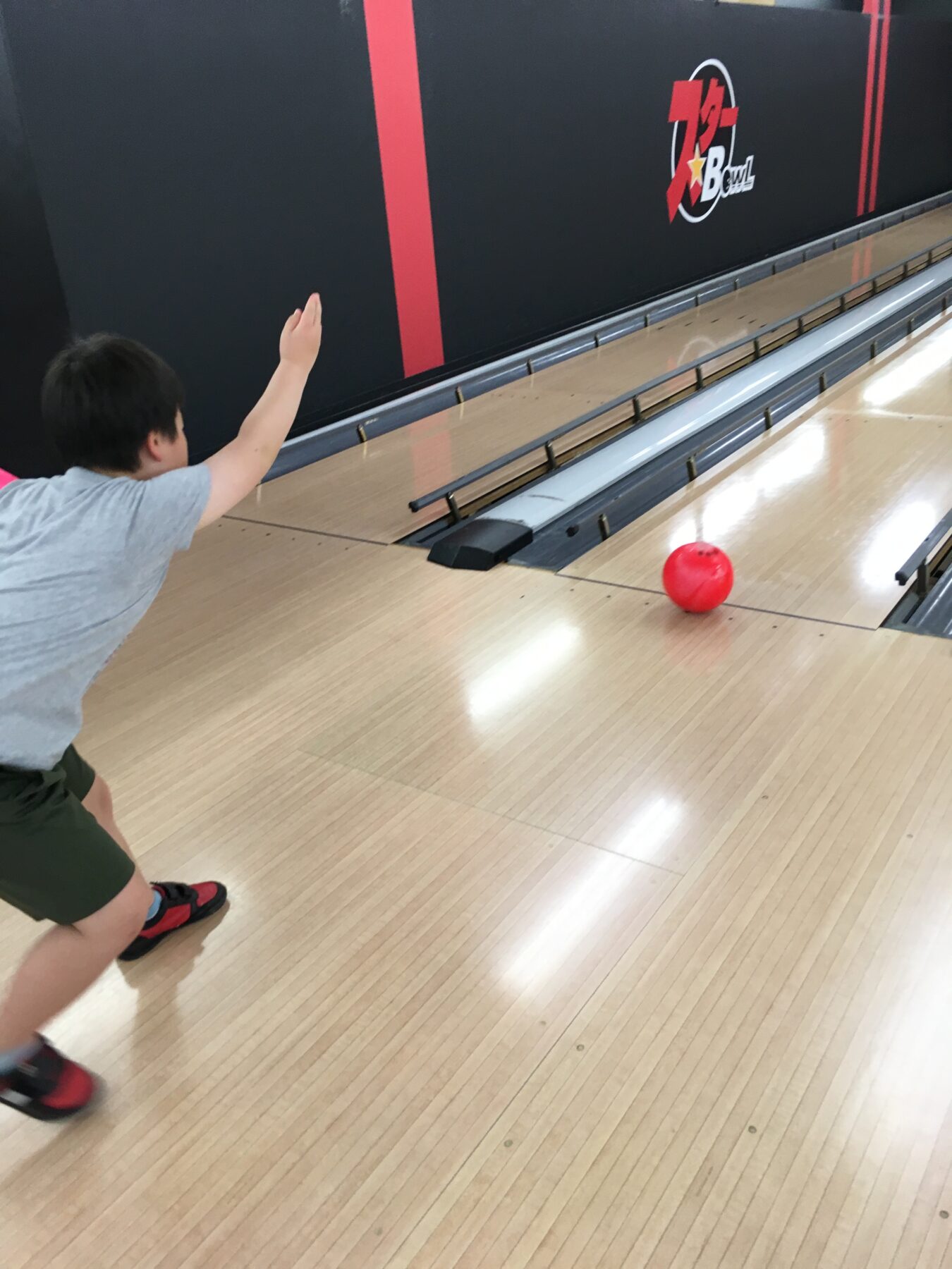 Child wearing gray shirt bowls a bright red ball down a wooden bowling lane toward the pins, in a bowling alley.