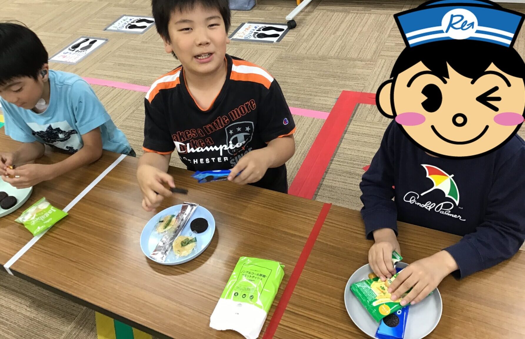 Three children sit at a table eating snacks; plates hold cookies and small snack packets on a classroom table with colored tape on the floor behind them.