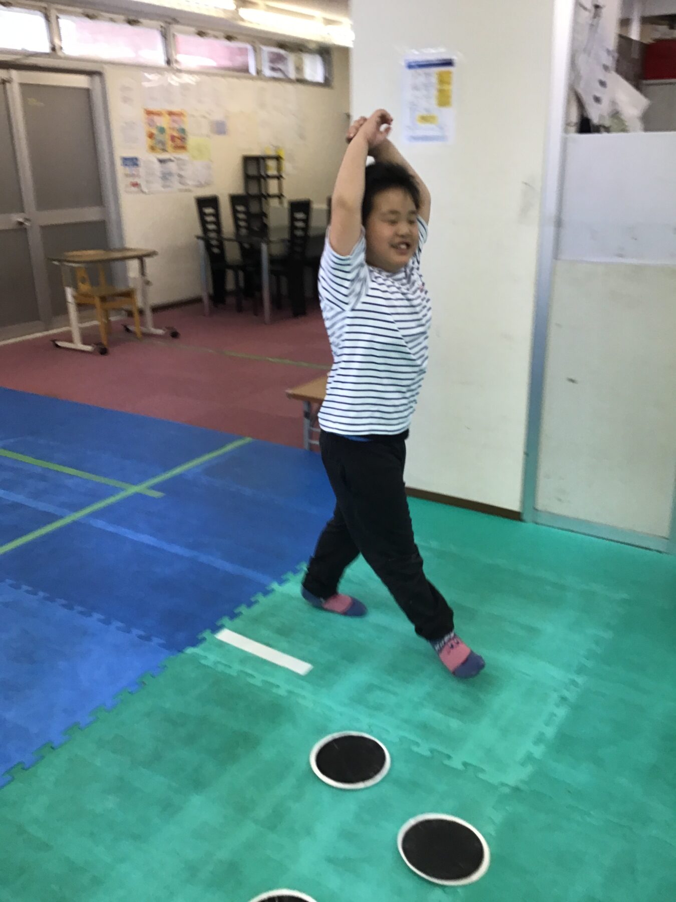 Child in a striped shirt jumps with arms raised on colorful foam mats in a gym or playroom.