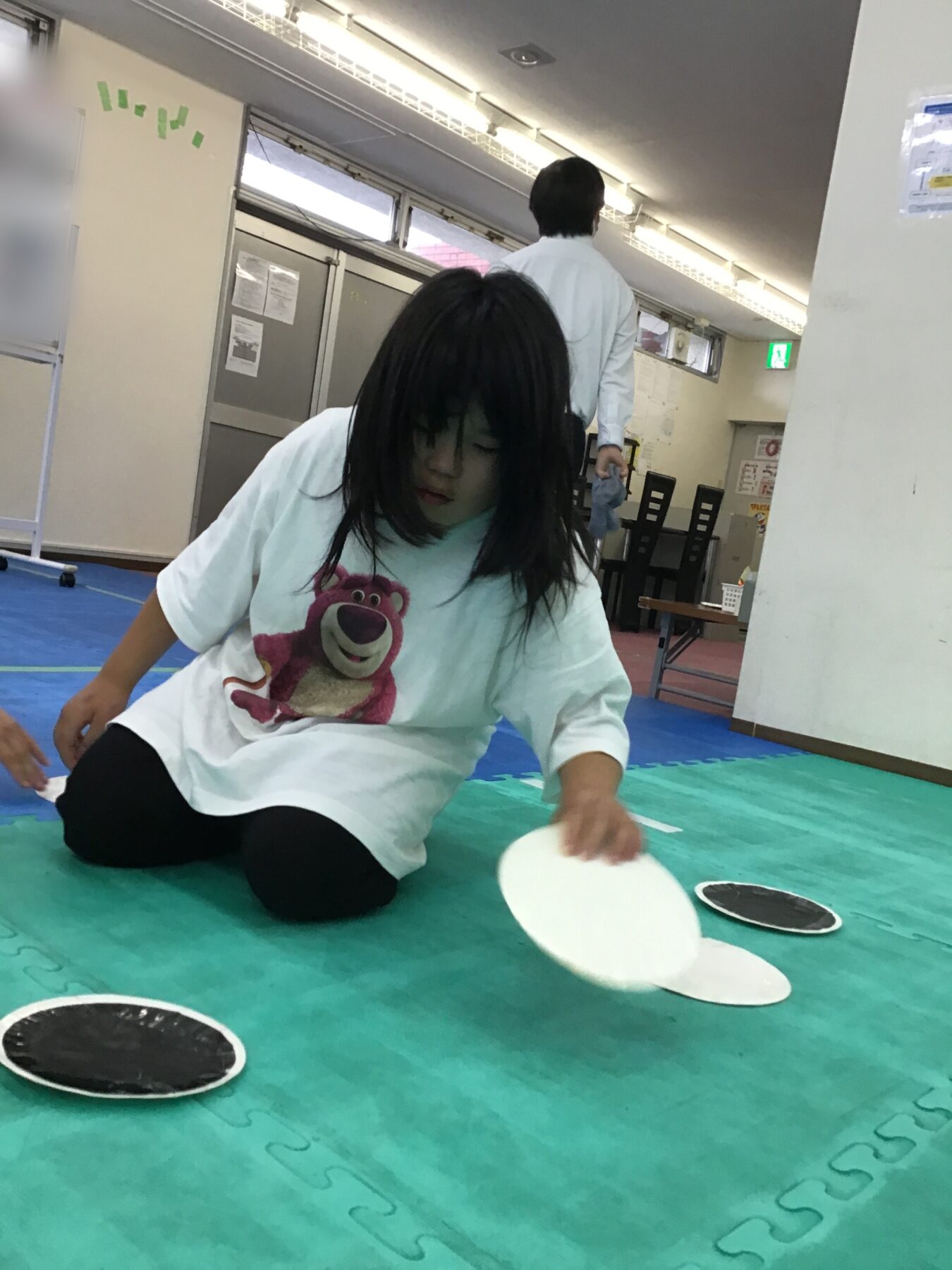Child in a pale blue shirt with a bear graphic sits on a green foam mat, reaching for white discs with black centers as another person walks in the background.