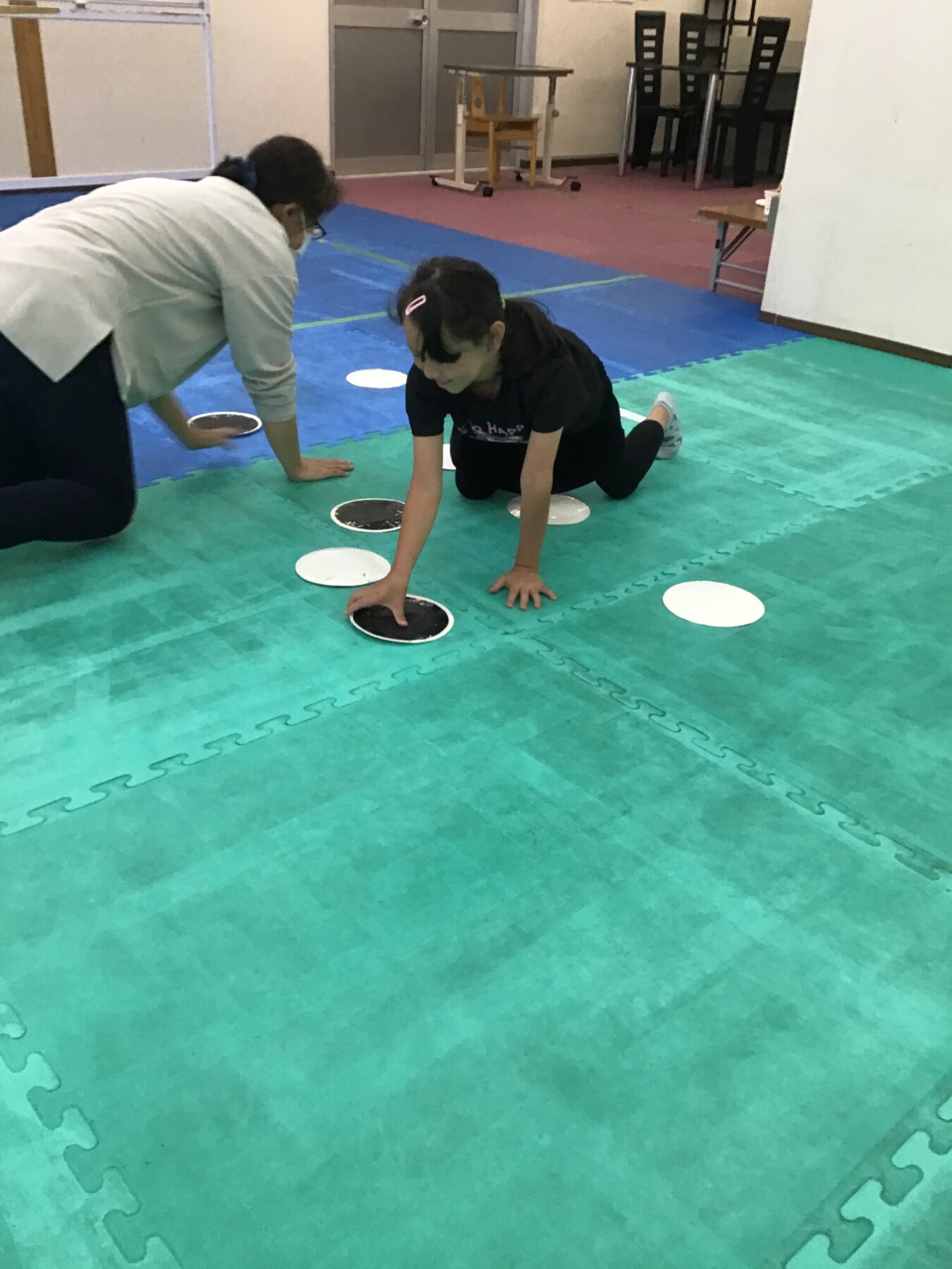 Young girl on hands and knees on a green foam mat, reaching for circular discs while an adult watches nearby in a gym-like room.
