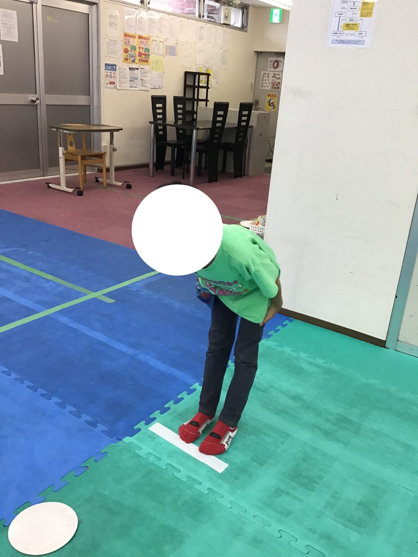 Child in a bright green shirt and red socks stands on a white floor line in a padded play area with blue and green mats and wall posters in the background.