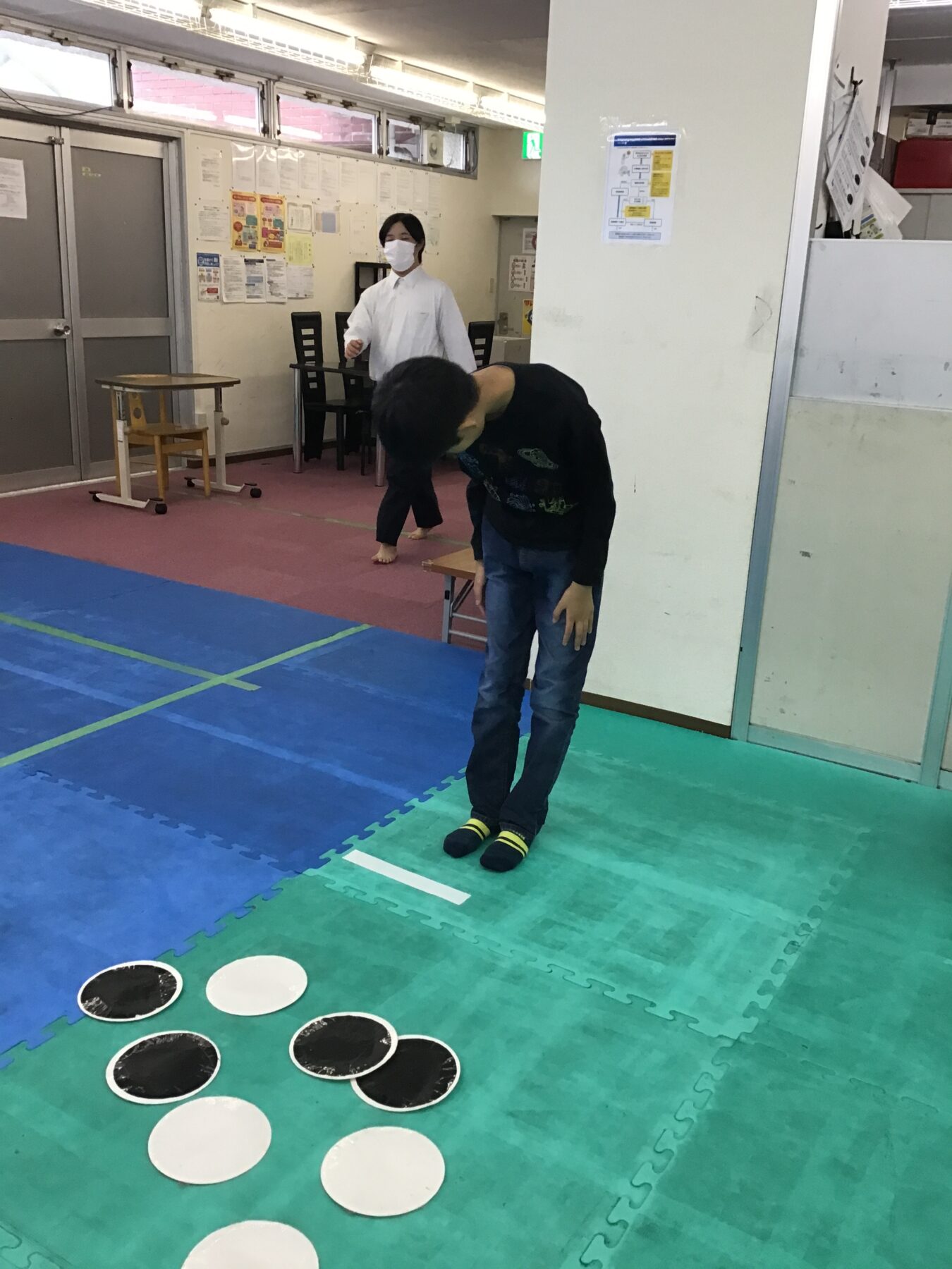 Boy in a black hoodie standing on a green mat, bending forward toward a line of white circles and black discs on the floor in a gym-like room. A person in a white shirt and mask stands in the background near a wall with posters.