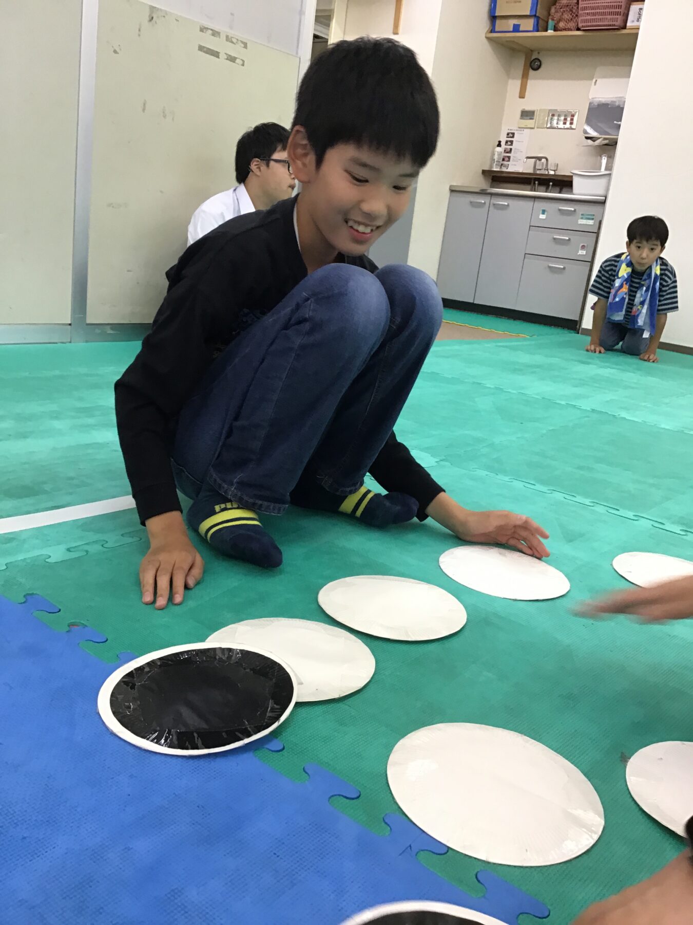 Smiling boy in a black shirt sits on a green foam mat, placing white circular discs on the floor; another child watches in the background in a classroom/kitchen area.