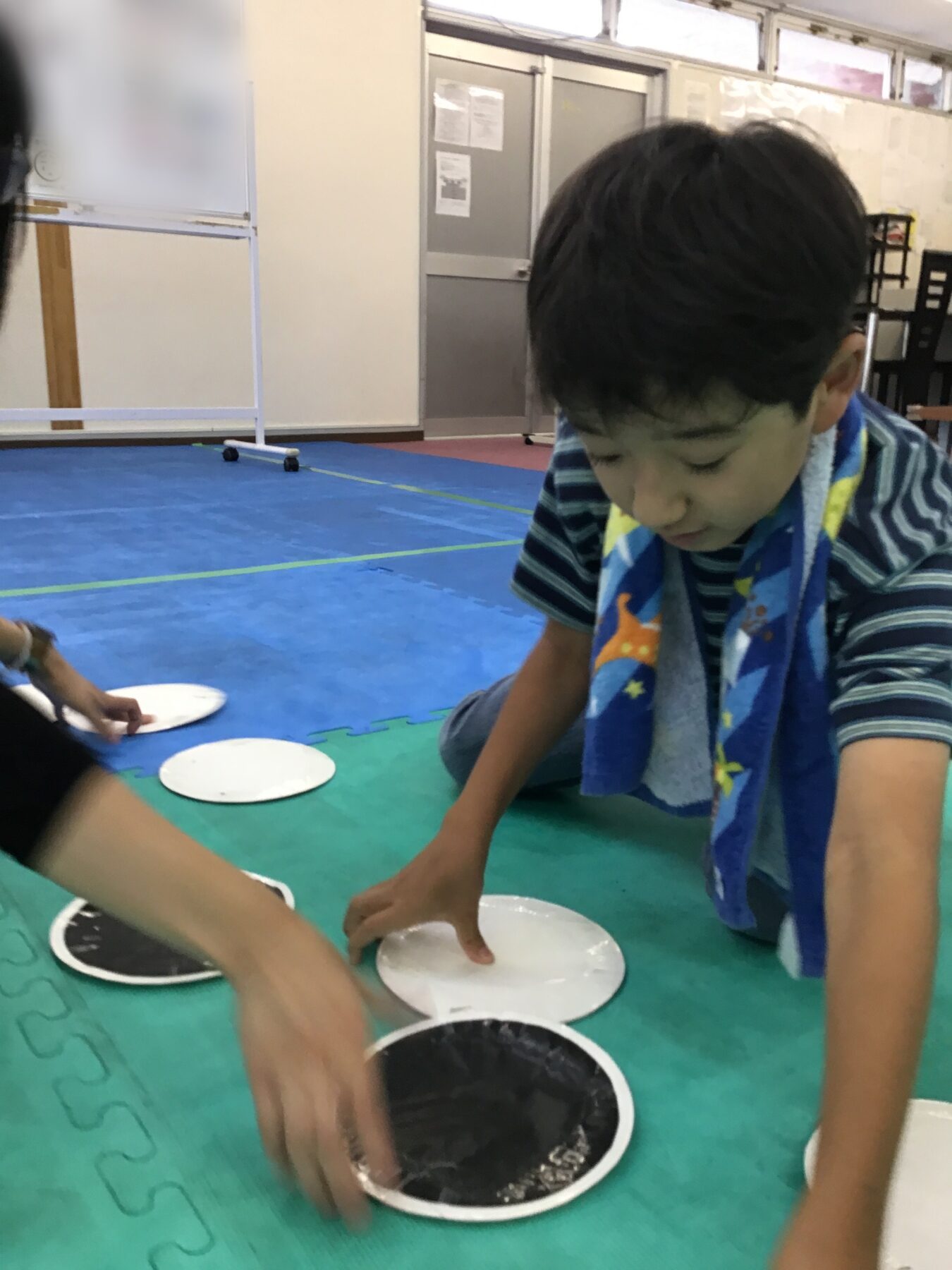 Two kids kneel at a low table, placing black and white discs on a green foam mat during a group activity in a gym.