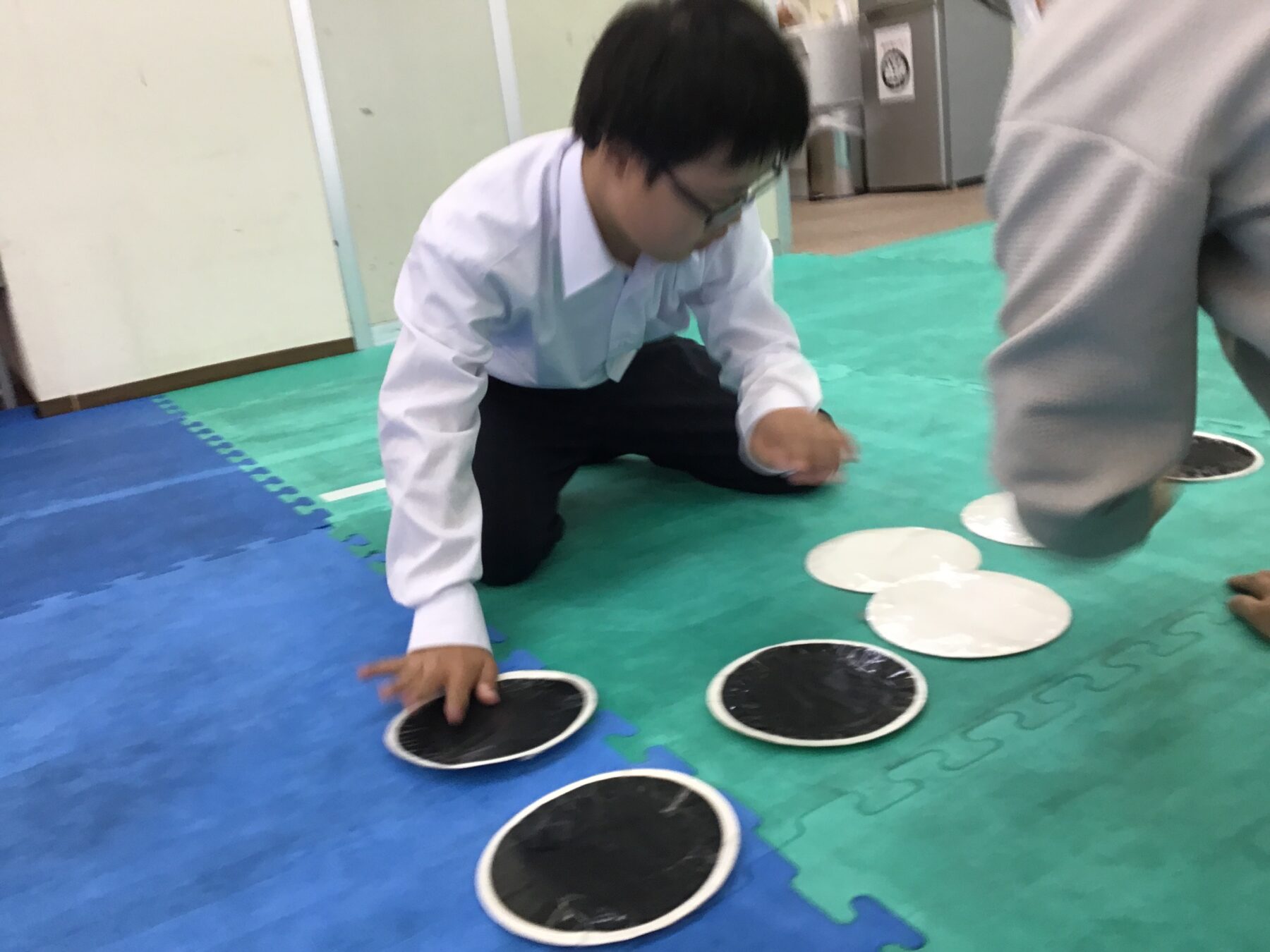Child kneeling on a green foam mat, arranging black circular discs as part of a floor game with white discs nearby.