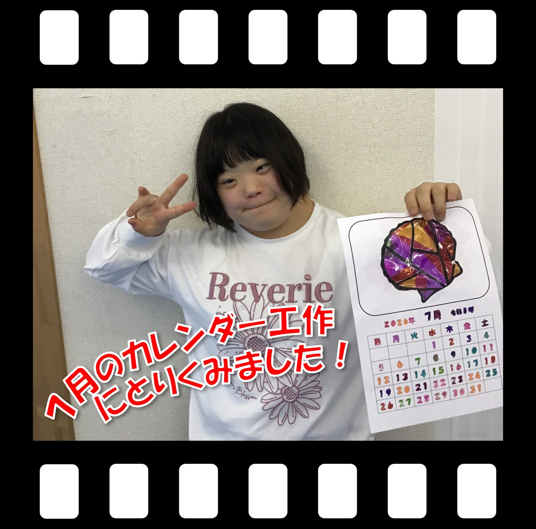 Child with short dark hair smiling and flashing a peace sign, holding a colorful calendar page against a light wall.
