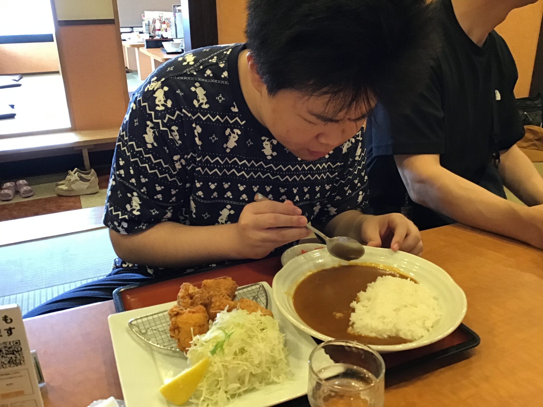 Person wearing a patterned shirt eats curry and rice at a table, with shredded cabbage, fried chicken pieces, and a lemon wedge on the side in a restaurant.