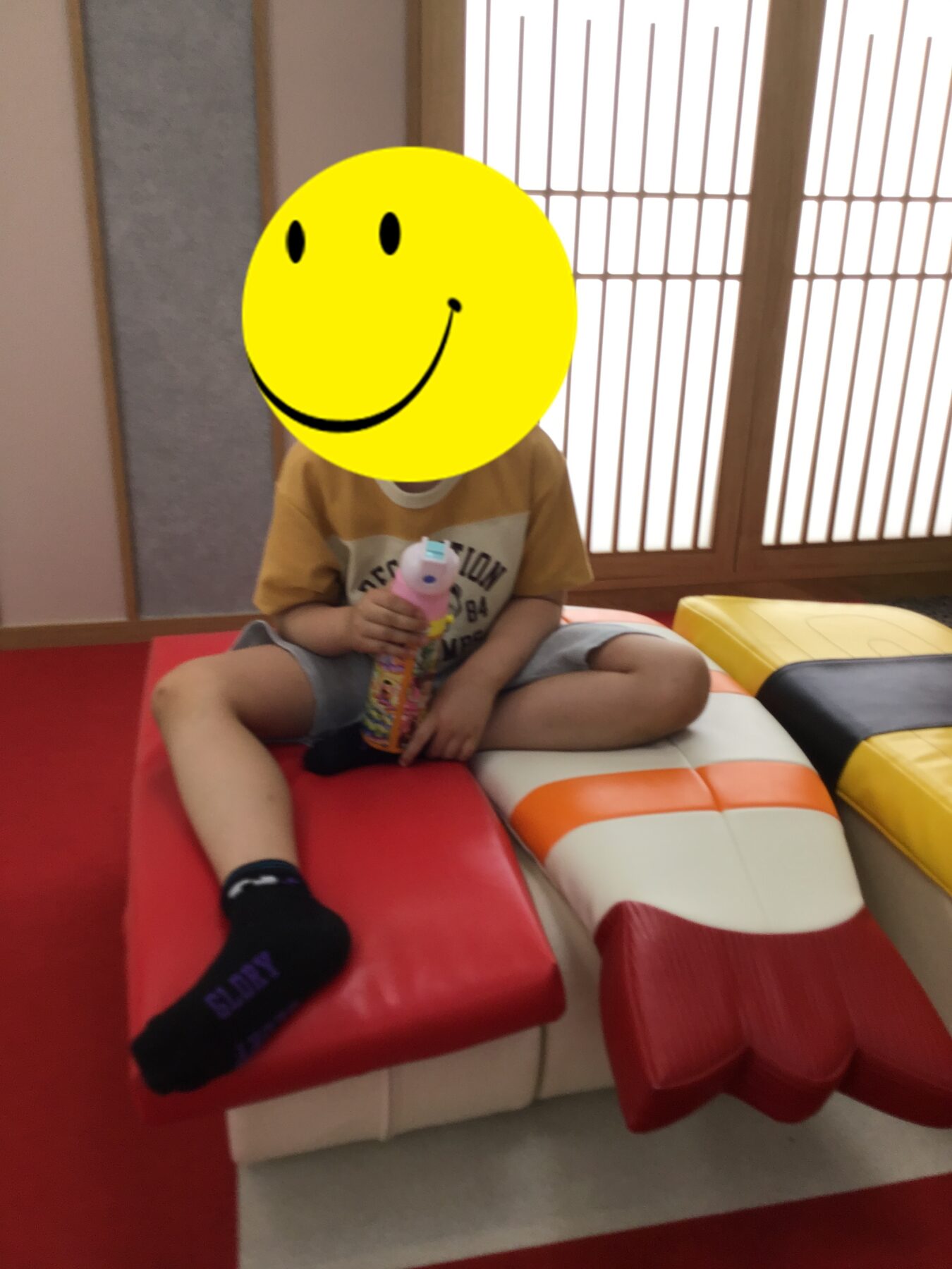 Child sitting cross‑legged on colorful foam seats, holding a bubble wand toy indoors. Faces covered by a yellow smiley overlay for privacy.