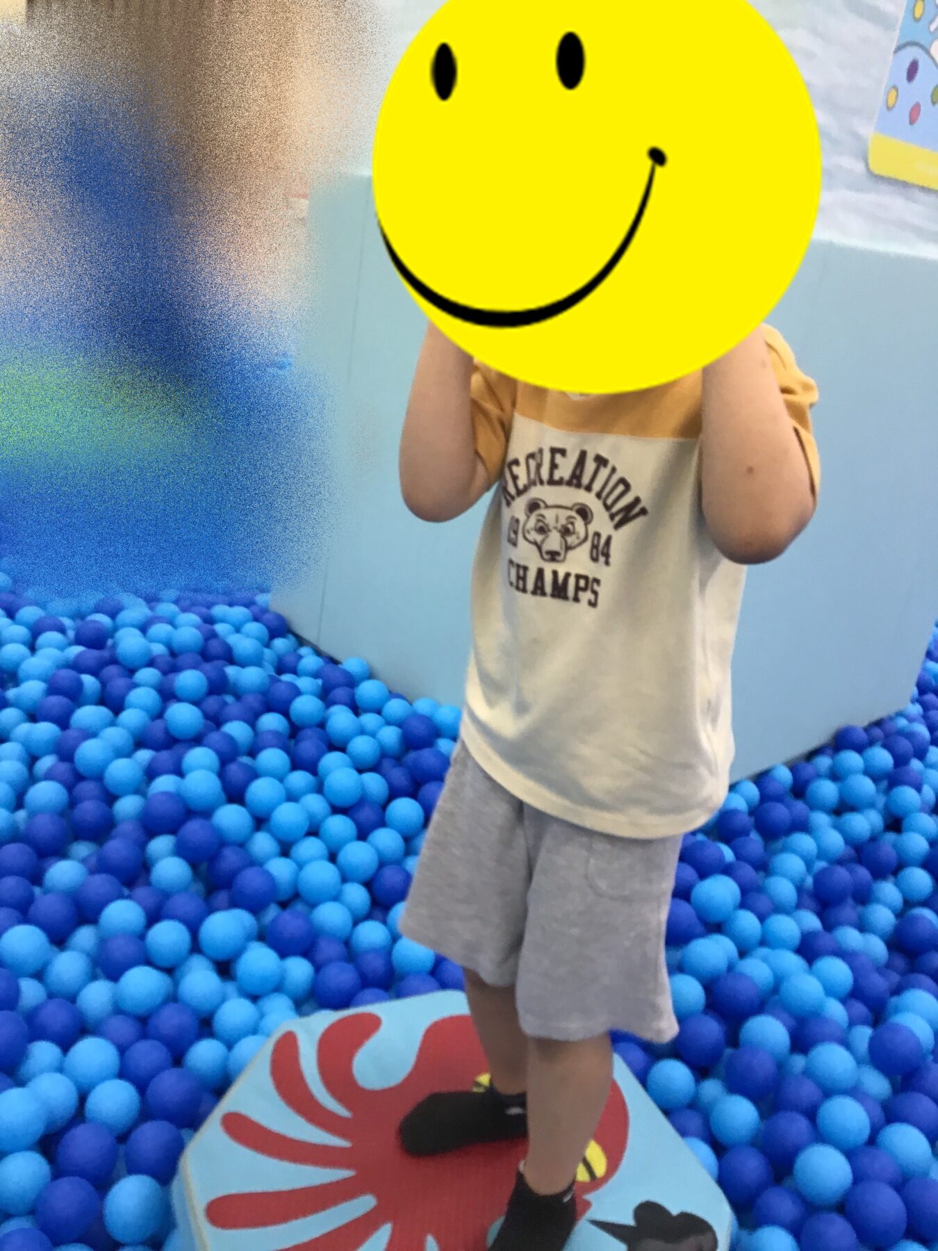 Child standing in a blue ball pit wearing a beige shirt and gray shorts; a large yellow smiley face covers their face for privacy. The child is on a colorful play mat with a red design nearby.