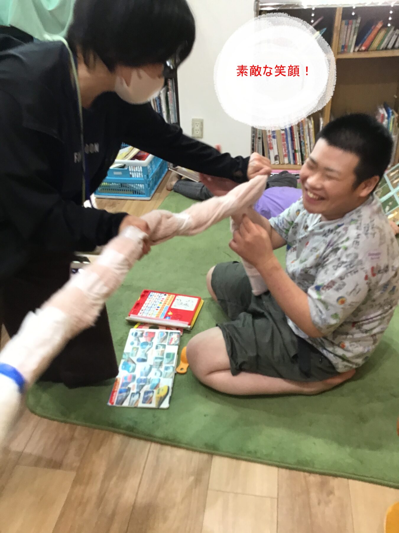 Man in a floral shirt sits on a green rug, smiling as a helper in black wraps his forearm with a bandage; bookshelves and toys in the background.