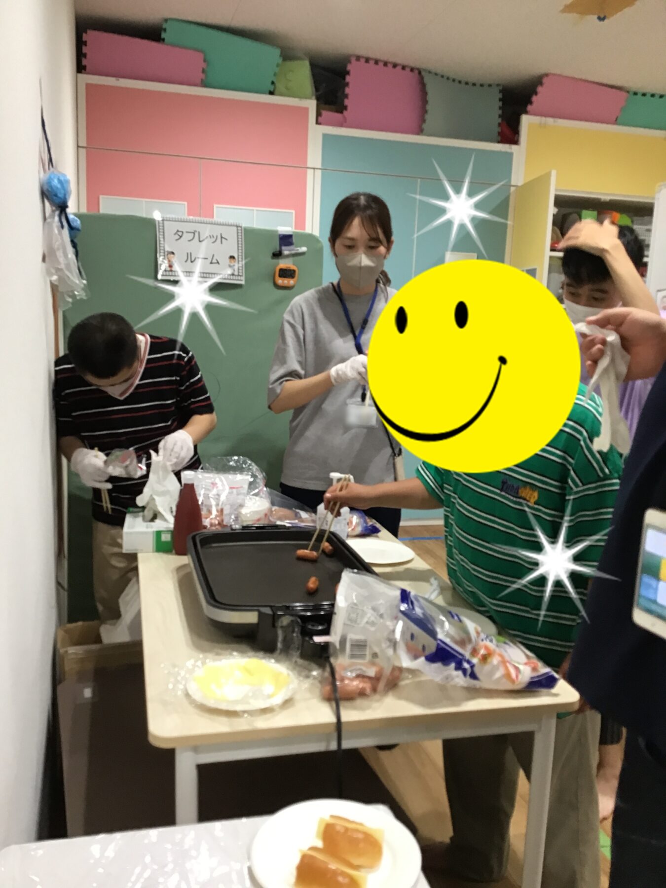 People wearing masks and gloves cooking at a tabletop grill in a colorful room; a large yellow smiley covers one person’s face.