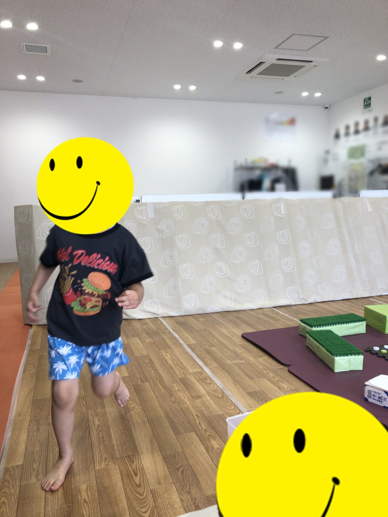 Child running barefoot in a bright indoor play area with foam mats and a large yellow smiley-faced figure obscuring their head.