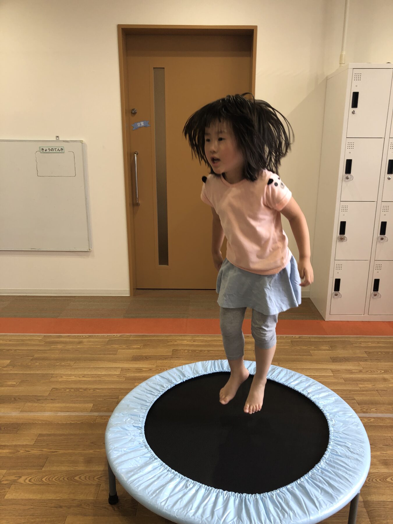 Young girl with dark hair, wearing a pink shirt and light blue skirt, jumping on a small blue-trimmed indoor trampoline in a room with lockers and a wooden floor.