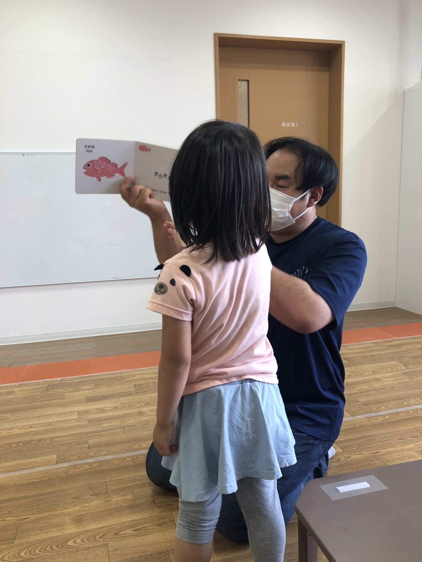 Man kneeling with a mask helps a young girl read a pink fish picture book in a classroom.
