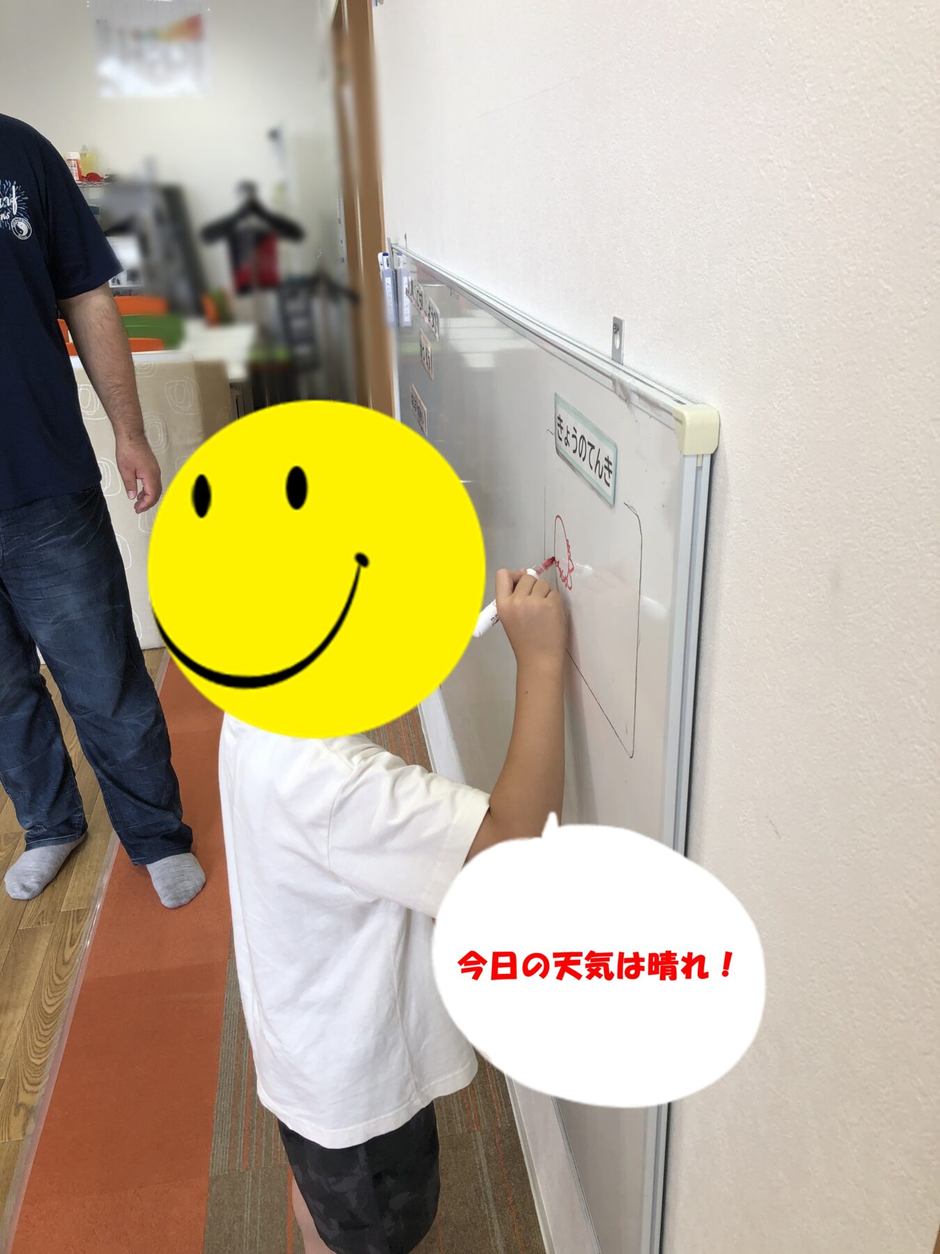 Child writes on a whiteboard mounted on a wall in a hallway; a large yellow smiley covers their face, and the board shows Japanese text in red reading '今日の天気は晴れ！'